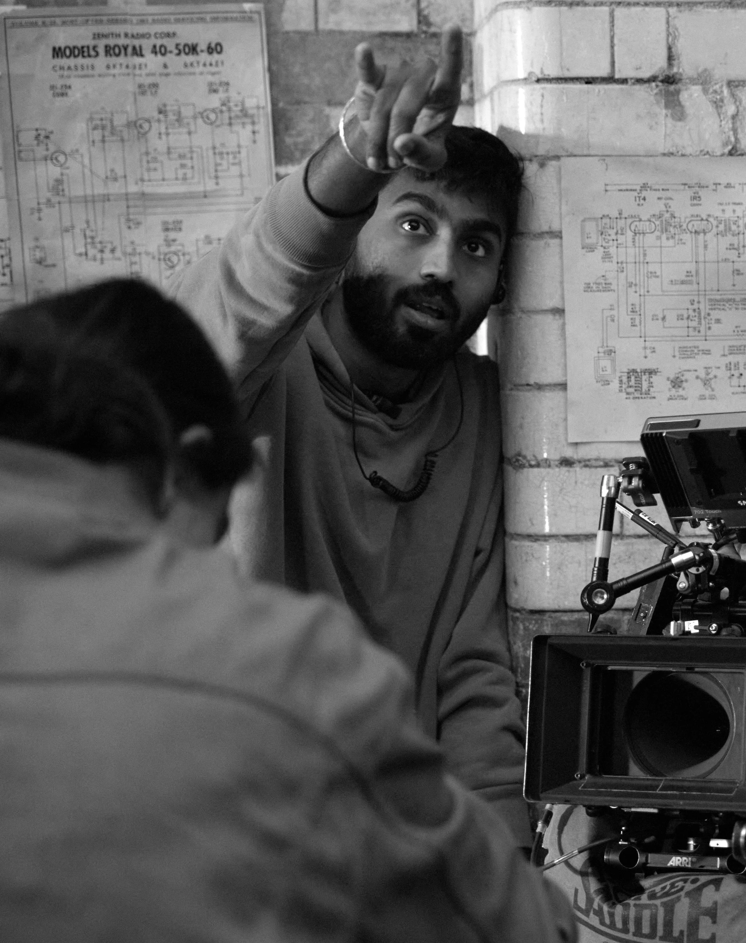 A man with a beard and short hair pointing upward, sitting on a chair next to a brick wall with architectural plans pinned on it. There is a person with braided hair in the foreground, with a camera setup nearby.
