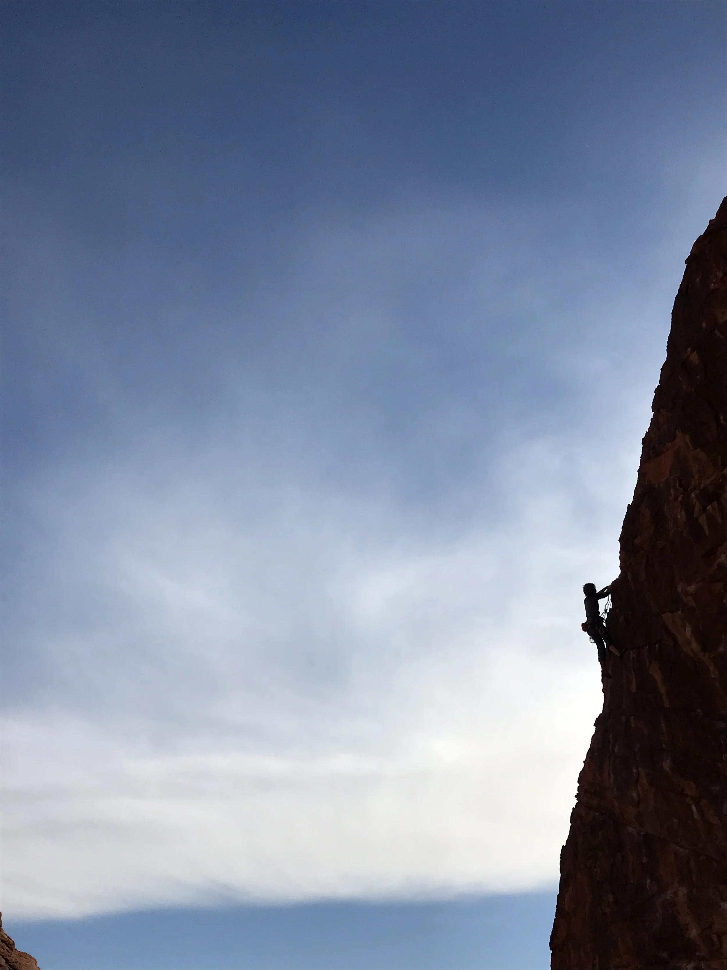 Silhouette of a person climbing a red rock cliff against a sky with clouds.