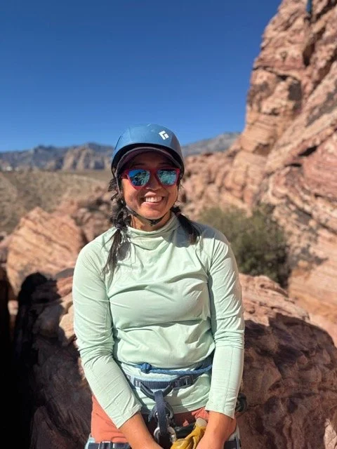 A woman wearing a light green long-sleeve shirt, sunglasses, and a helmet, smiling outdoors near rocky terrain with mountains in the background.