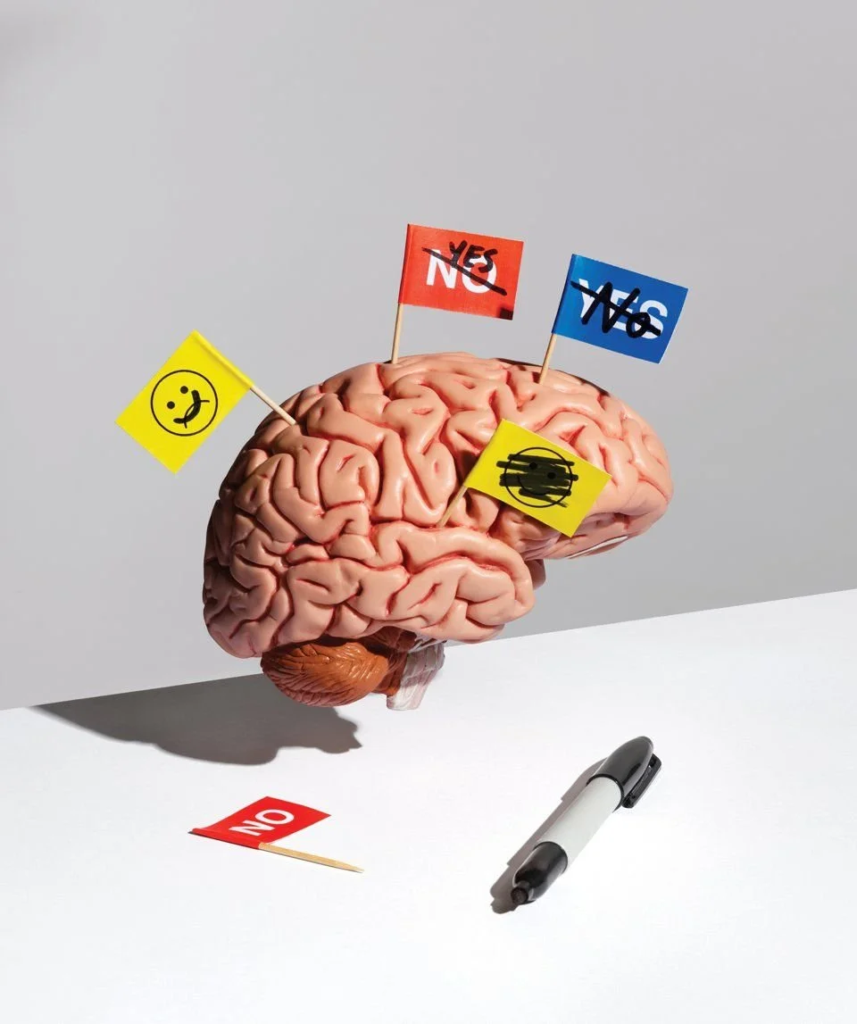 A human brain with small protest signs attached, including a yellow smiley face, a red 'NO' sign, a blue 'YES' sign, and a yellow sign with a face wearing a mask. There is also a 'NO' sign on a toothpick on the table and a black marker nearby. The background is plain, with the brain casting a shadow.