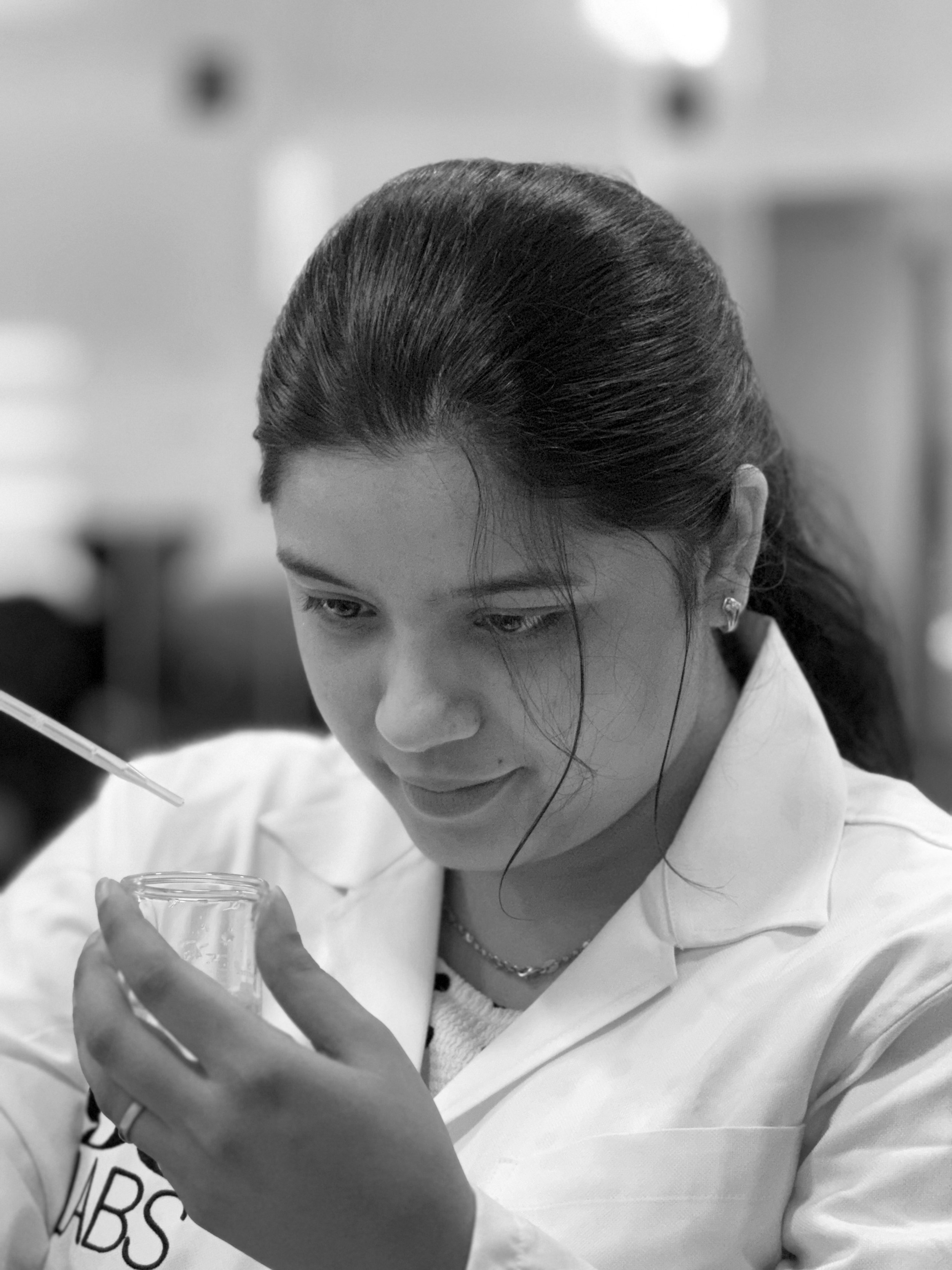 Black and white photo of a young woman in a lab coat, examining a small test tube in her left hand while using a pipette with her right hand.