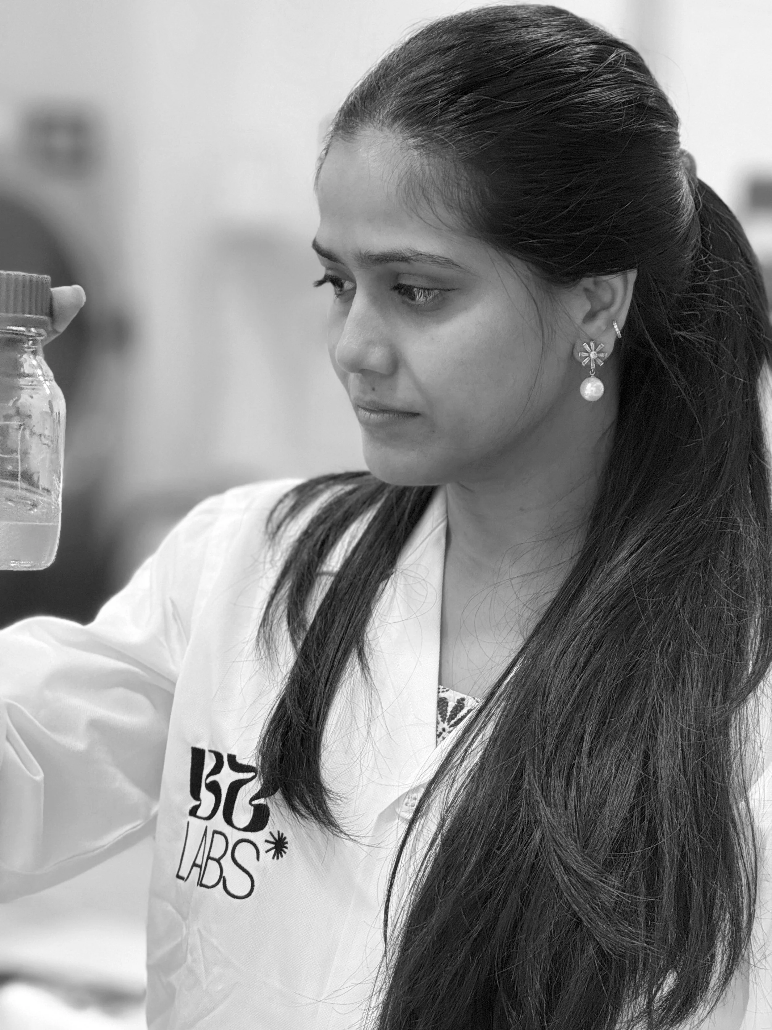 A woman with long dark hair, wearing earrings and a white lab coat with the logo 'BC LABS,' is holding a small jar and looking at it with concentration in a laboratory setting.