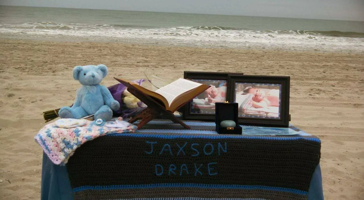 A memorial setup on the beach with a teddy bear, framed photos, an open book on a stand, and a box necklace, with the names 'JAXSON' and 'DRAKE' knitted into a blanket.