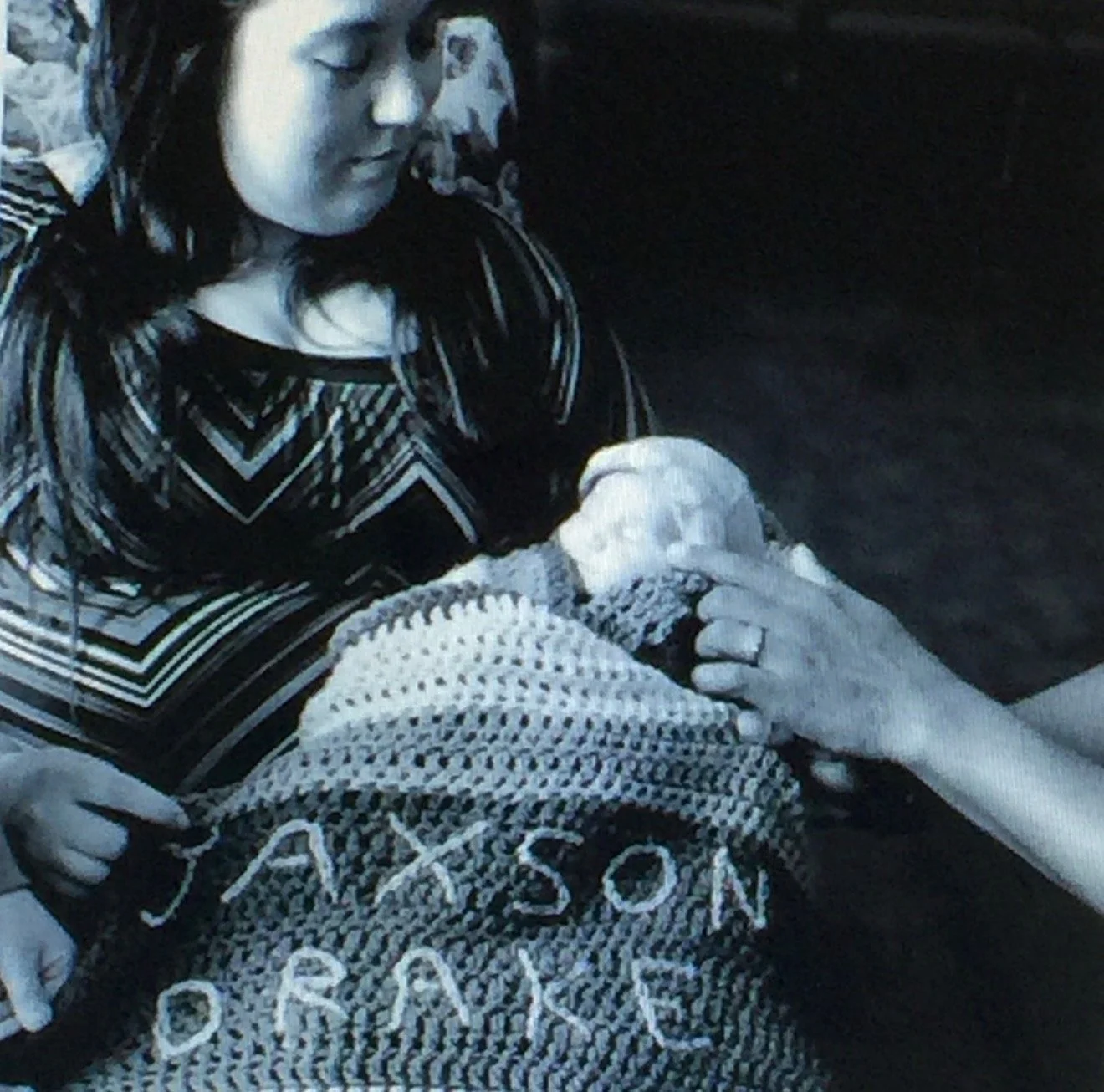 A woman with dark hair holds a baby wrapped in a blanket with the words "JESUS LOVE" embroidered on it. The woman appears to be kneeling or sitting, with her eyes closed, in a peaceful moment.