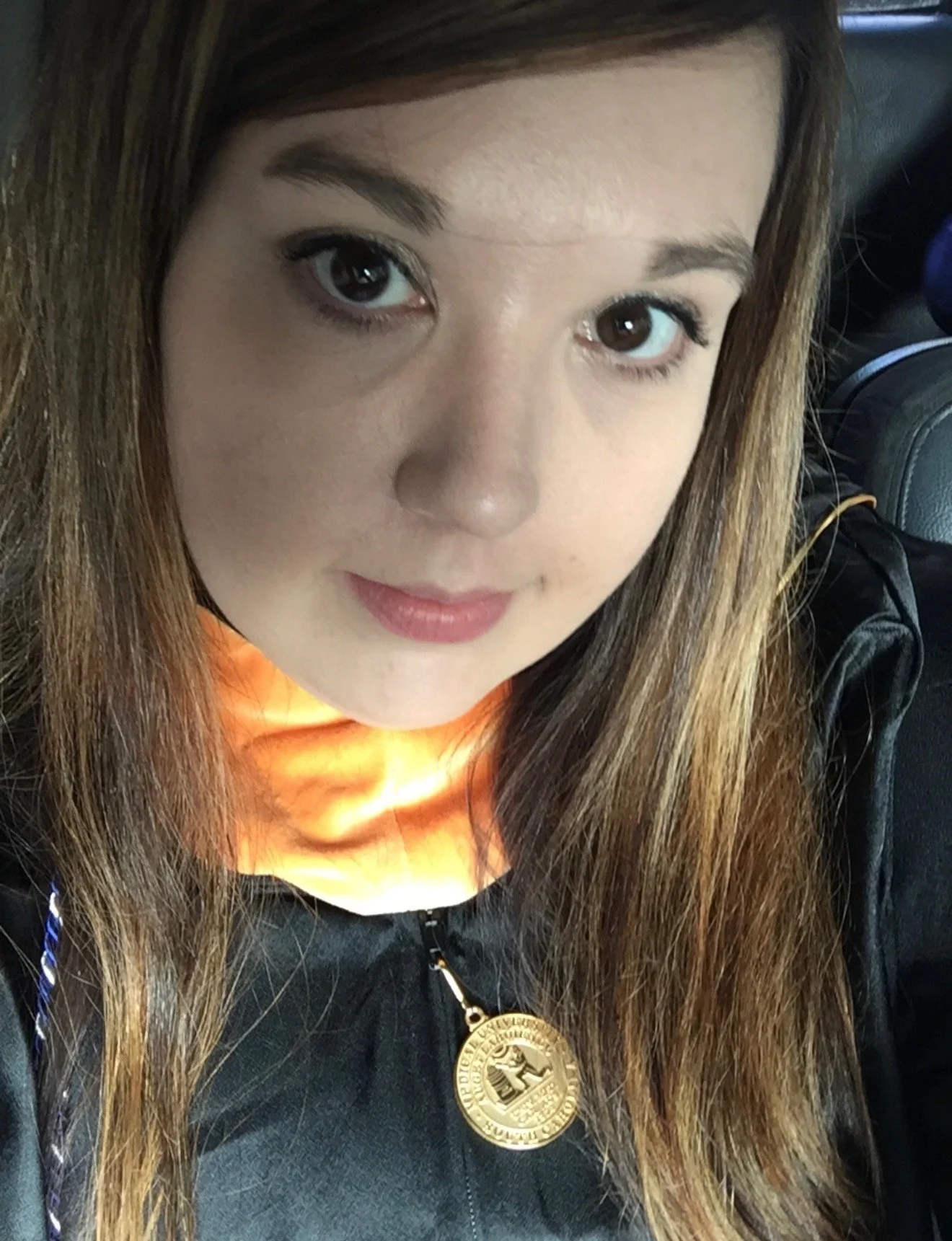Close-up of a woman with long brown hair, wearing makeup, and a black jacket with a gold medal-like pendant around her neck, sitting in a vehicle.