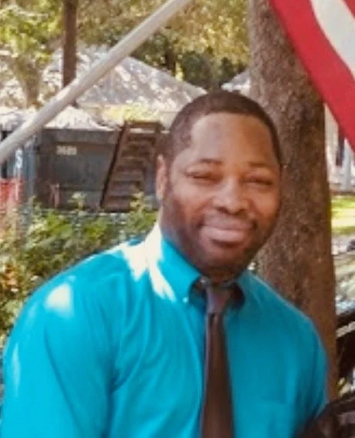 A man with short hair and a goatee, wearing a bright blue shirt and a black tie, standing outdoors near a tree with an American flag partially visible in the background.