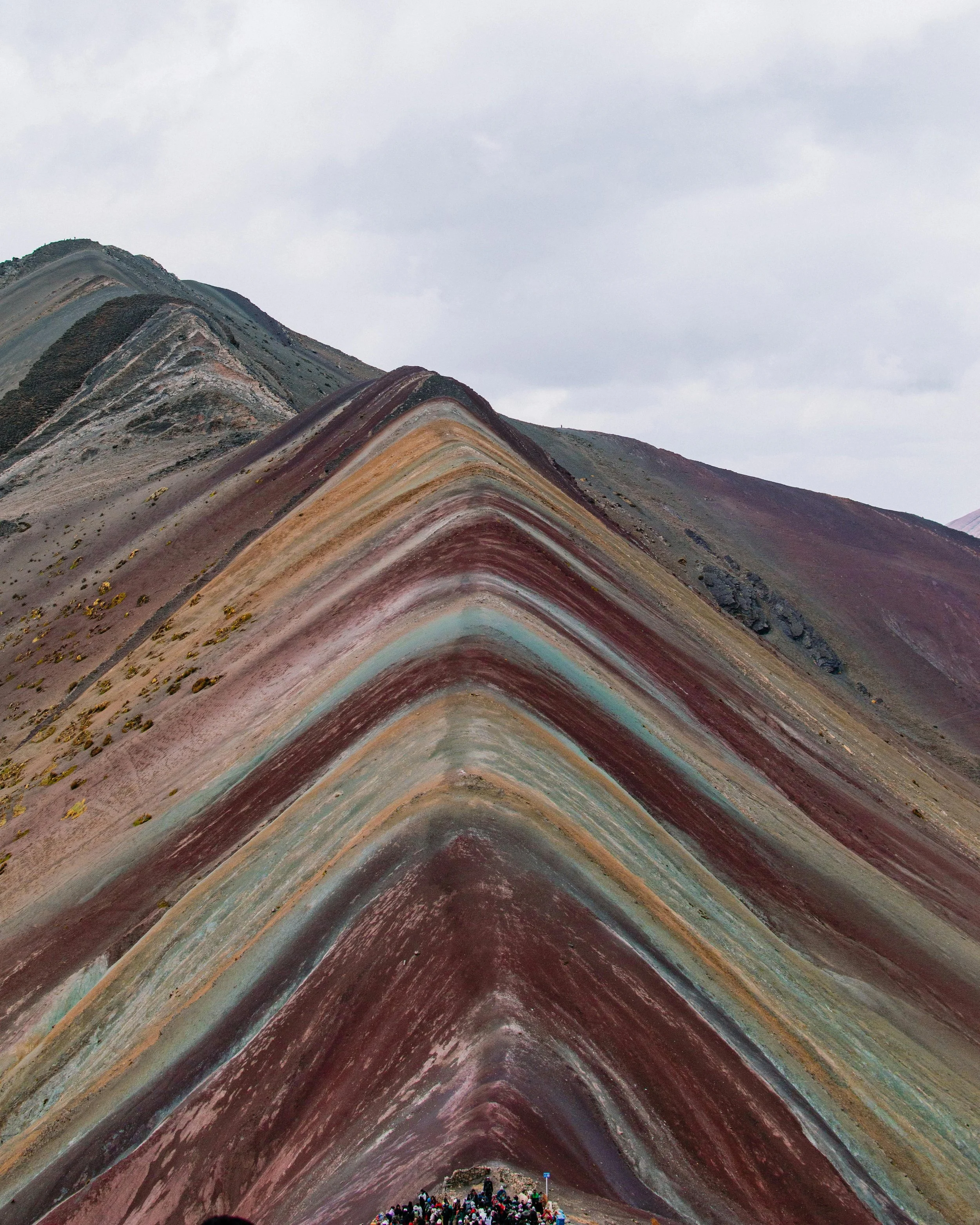 Colorful, striped mountain formation with a large group of people at the base, cloudy sky above.