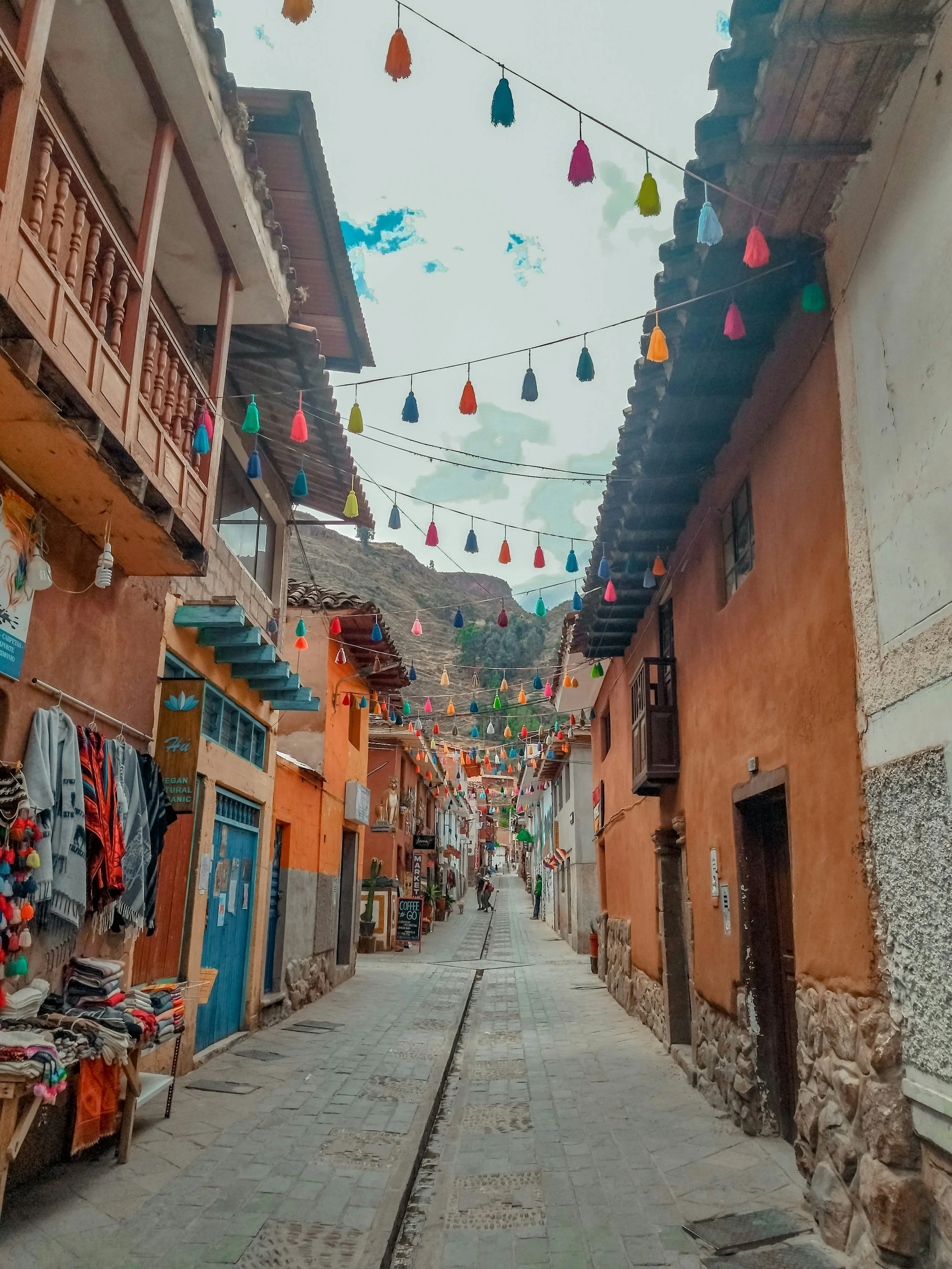 A narrow cobblestone street in a colorful village with hanging decorative tassel lights, shops on both sides selling clothing and souvenirs, and mountains in the background under a partly cloudy sky.