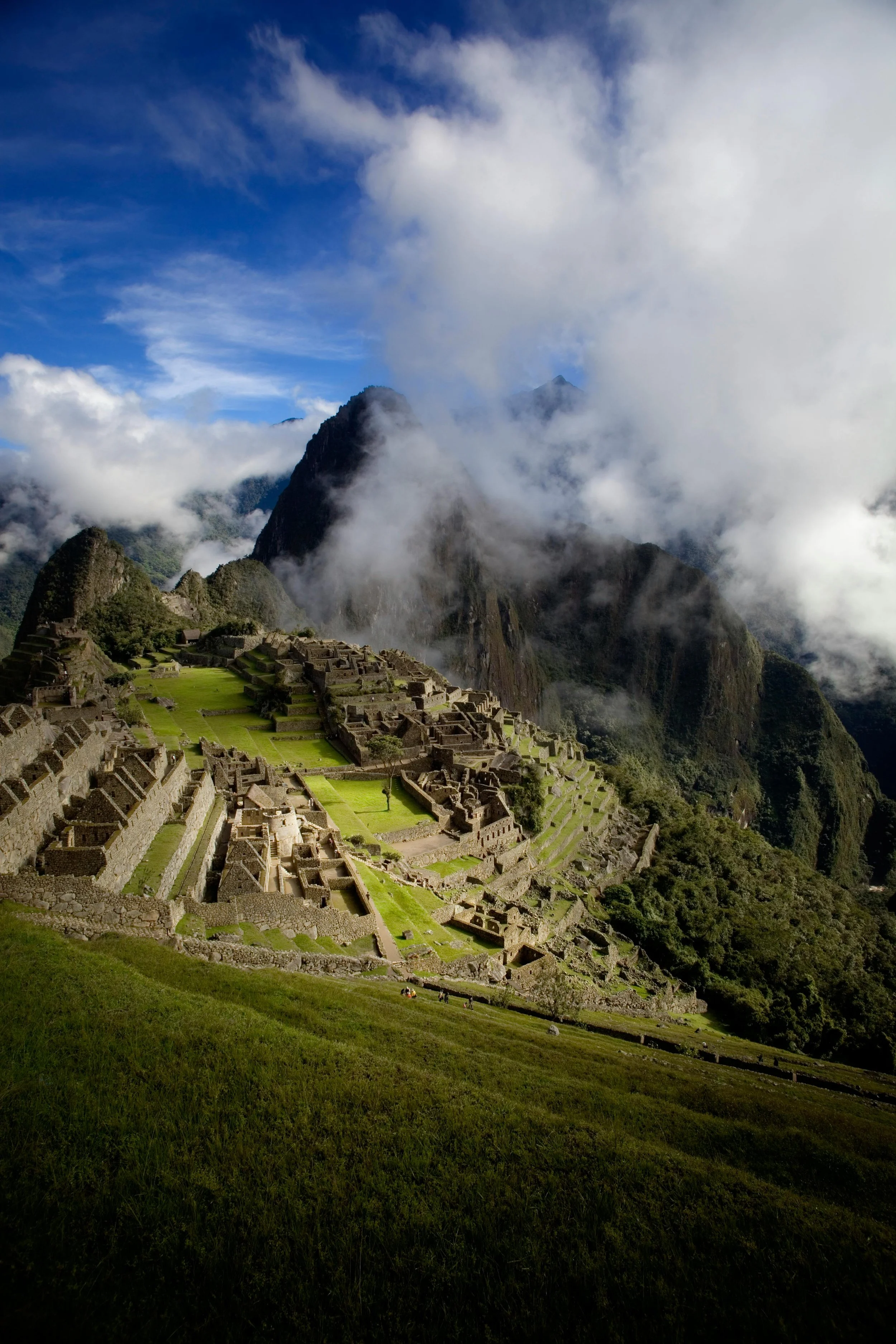 Machu Picchu, ancient Incan city, surrounded by mountains and clouds, with terraced slopes and stone structures.