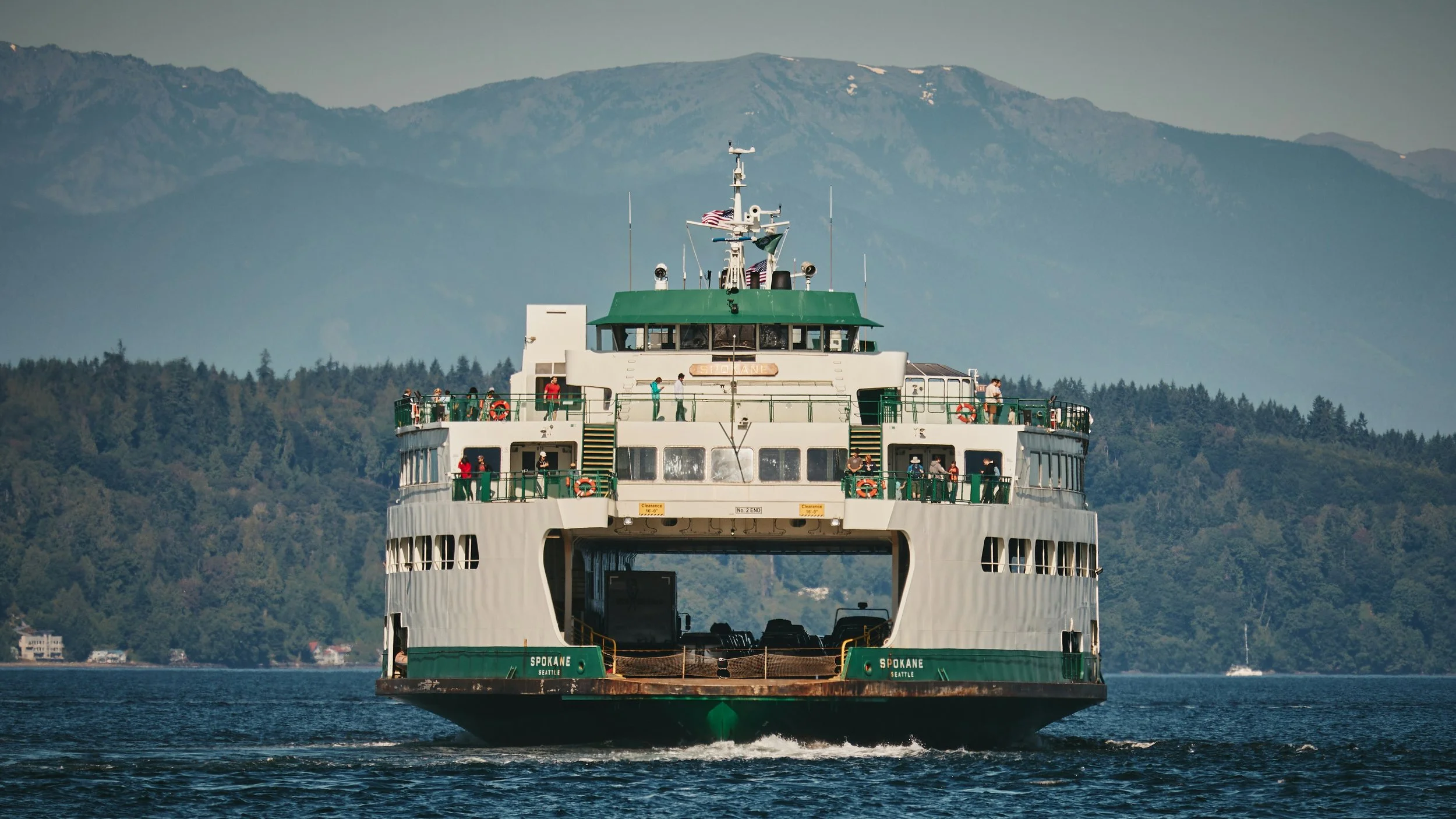 A large ferry boat with passengers onboard, sailing on a body of water with a backdrop of green mountainous terrain.