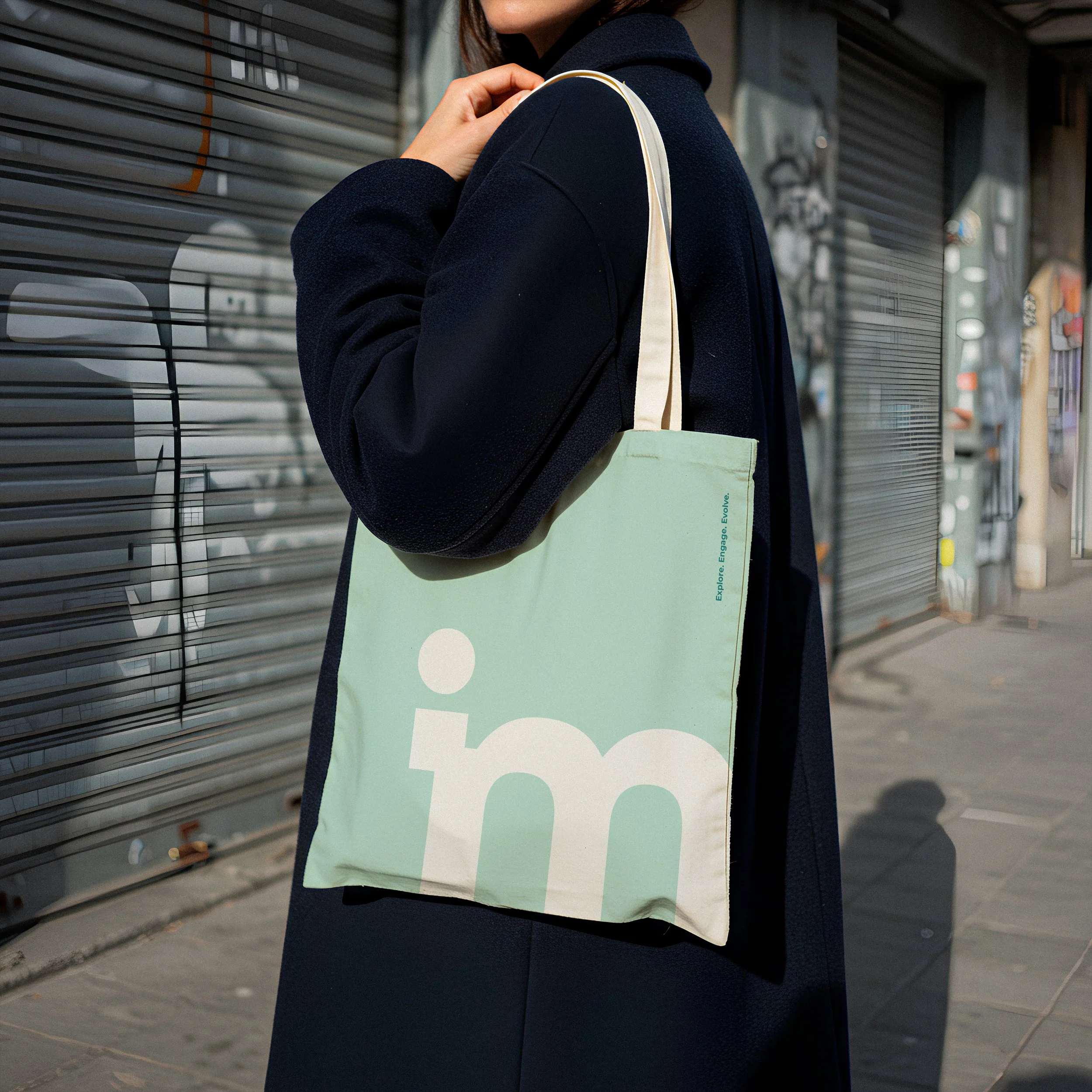 Person wearing a dark coat carrying a light green tote bag, standing next to metallic shop shutters in an urban setting.