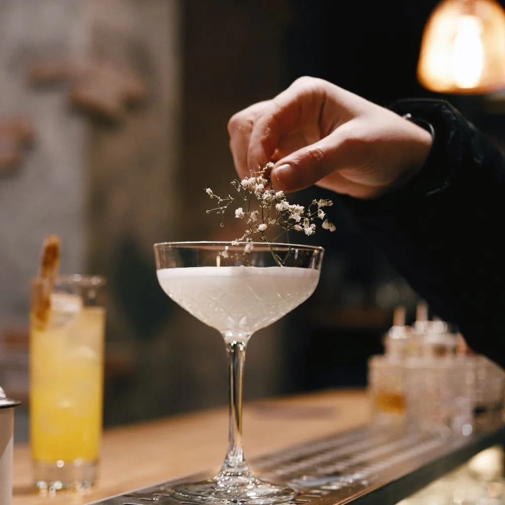 Person garnishing a cocktail with small white flowers in a coupe glass