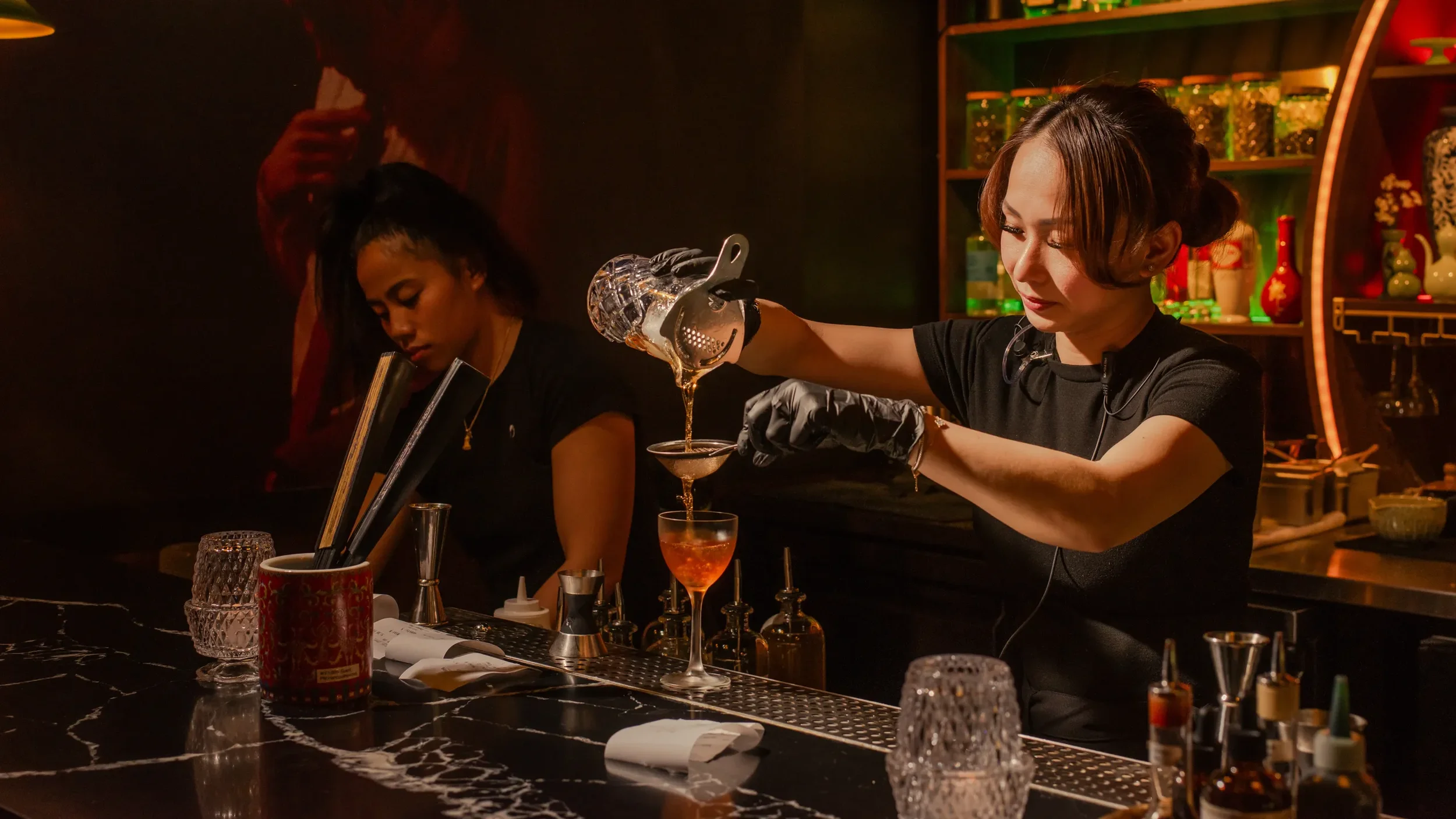 Two women working behind a bar, one pouring a cocktail through a strainer into a glass, the other leaning forward and working with bar tools; dimly lit with shelves of bottles and decorative items in the background.