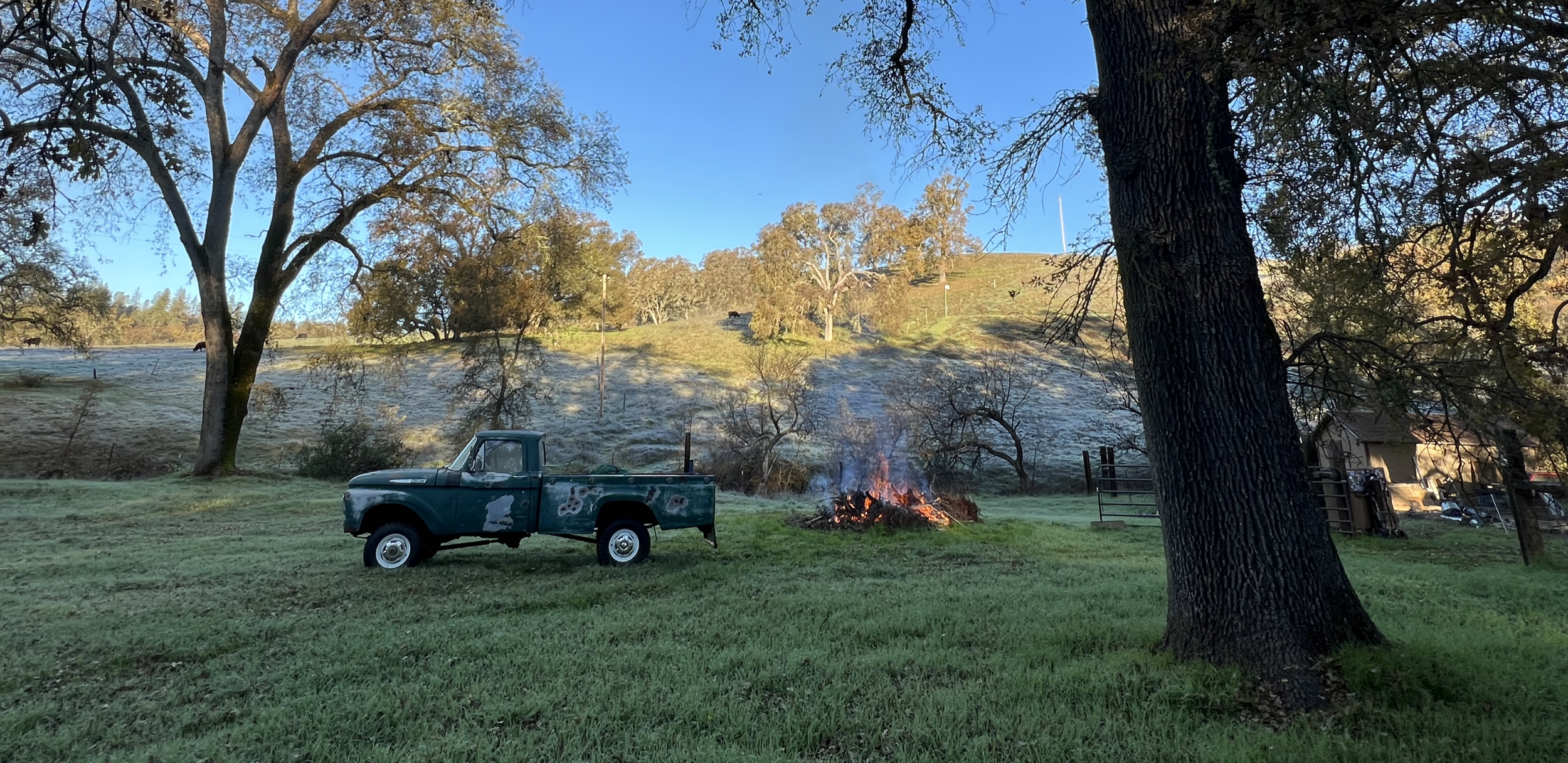 A rural scene with a green pickup truck parked on a grassy field, a small burning fire nearby, large trees casting shade, and a hillside in the background with more trees and a clear blue sky.