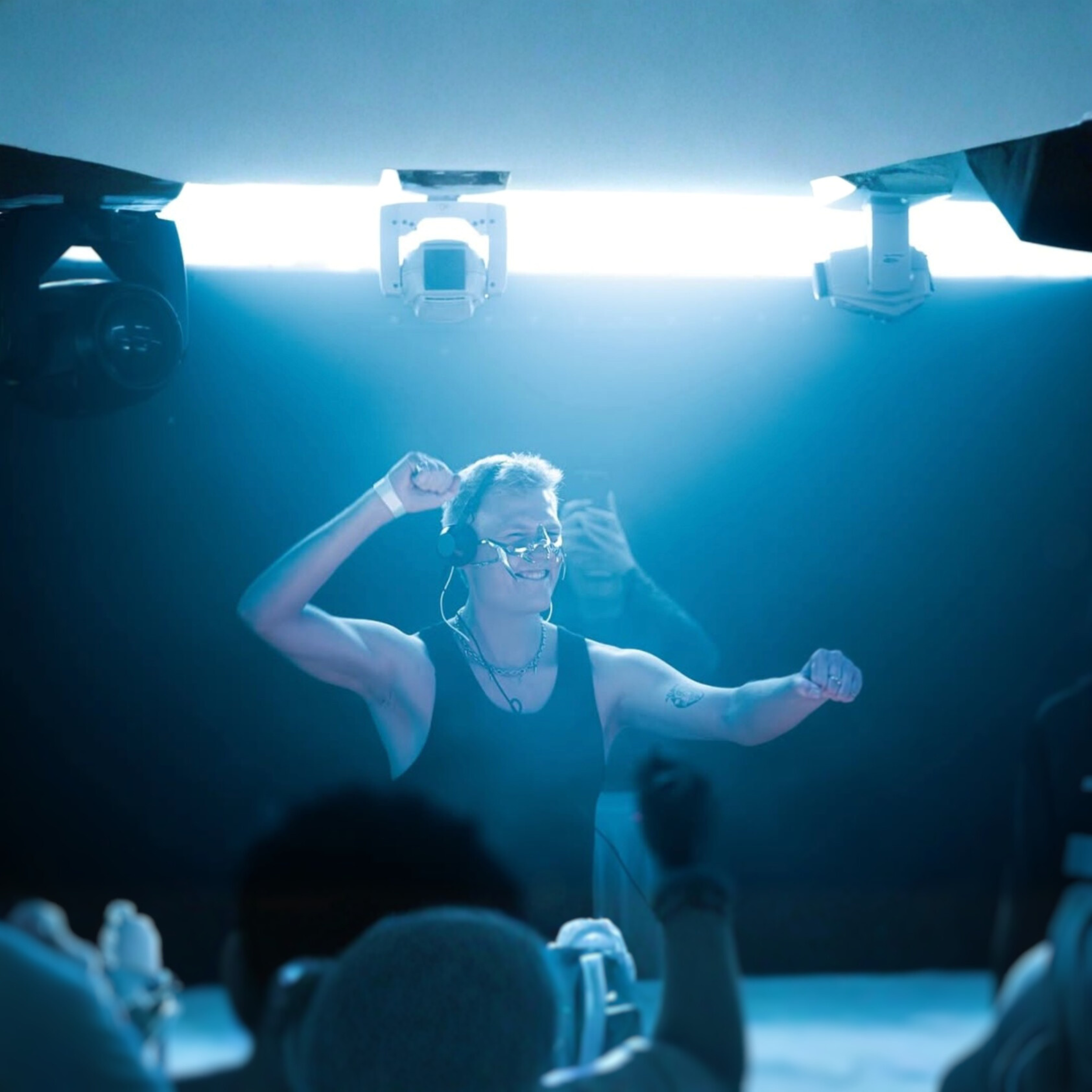 Eine Person mit Headset, erfrischt und lachend, posiert mit einem sportlichen Gruß, während sie in einem dunklen Raum mit blauer Beleuchtung auf einer Bühne ist. Das Publikum ist im Vordergrund zu sehen.