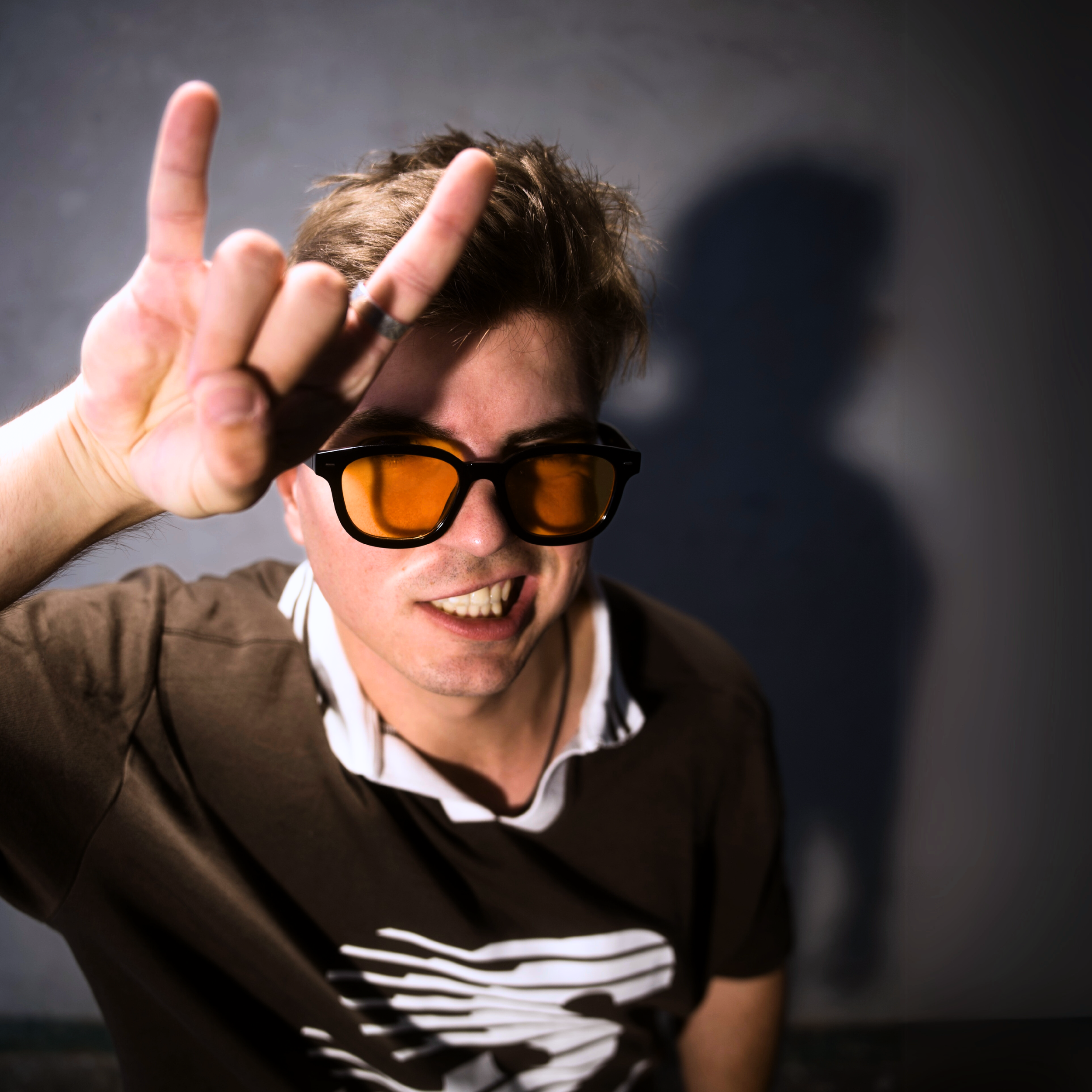 Junger Mann trägt Sonnenbrille, macht das Rockzeichen mit der Hand und lächelt.