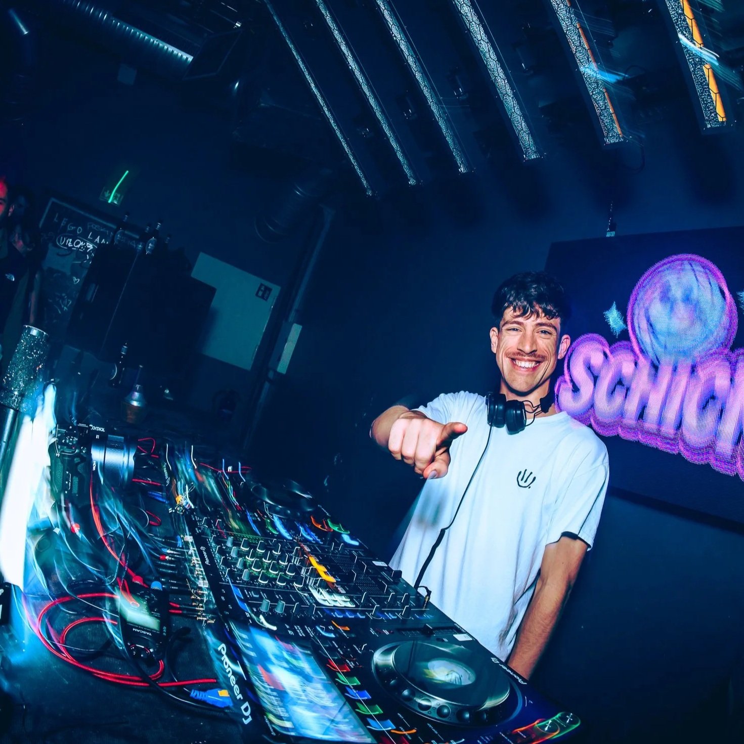 Ein junger Mann in einem weißen T-Shirt mit Kopfhörern um den Hals, der an einem DJ-Controller steht und zur Kamera lächelt, in einer dunklen Club-Umgebung mit bunten Neonlichtern im Hintergrund.