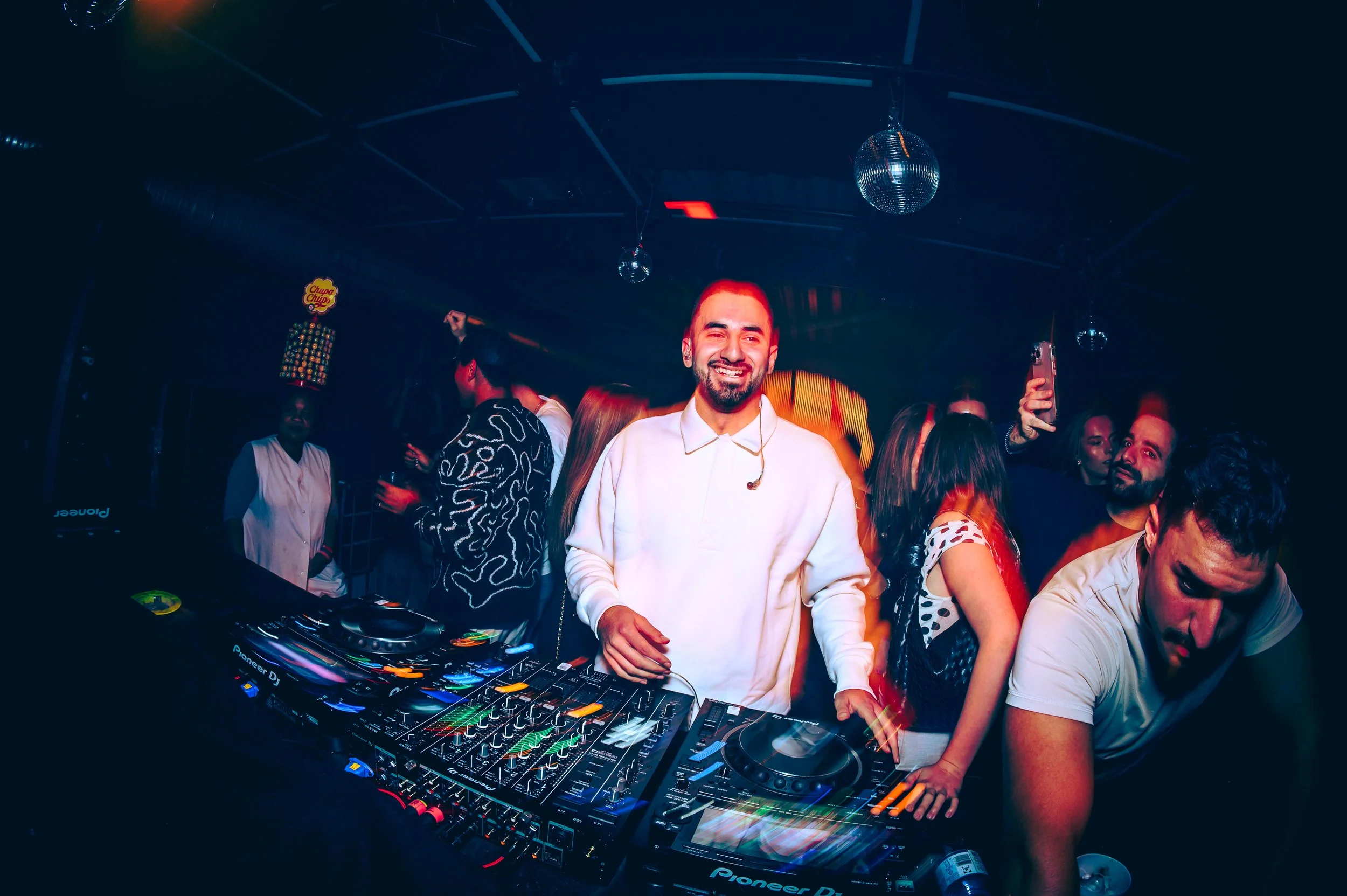 DJ in einem Club, umgeben von tanzenden Menschen, bunte Beleuchtung, Discokugeln an der Decke.