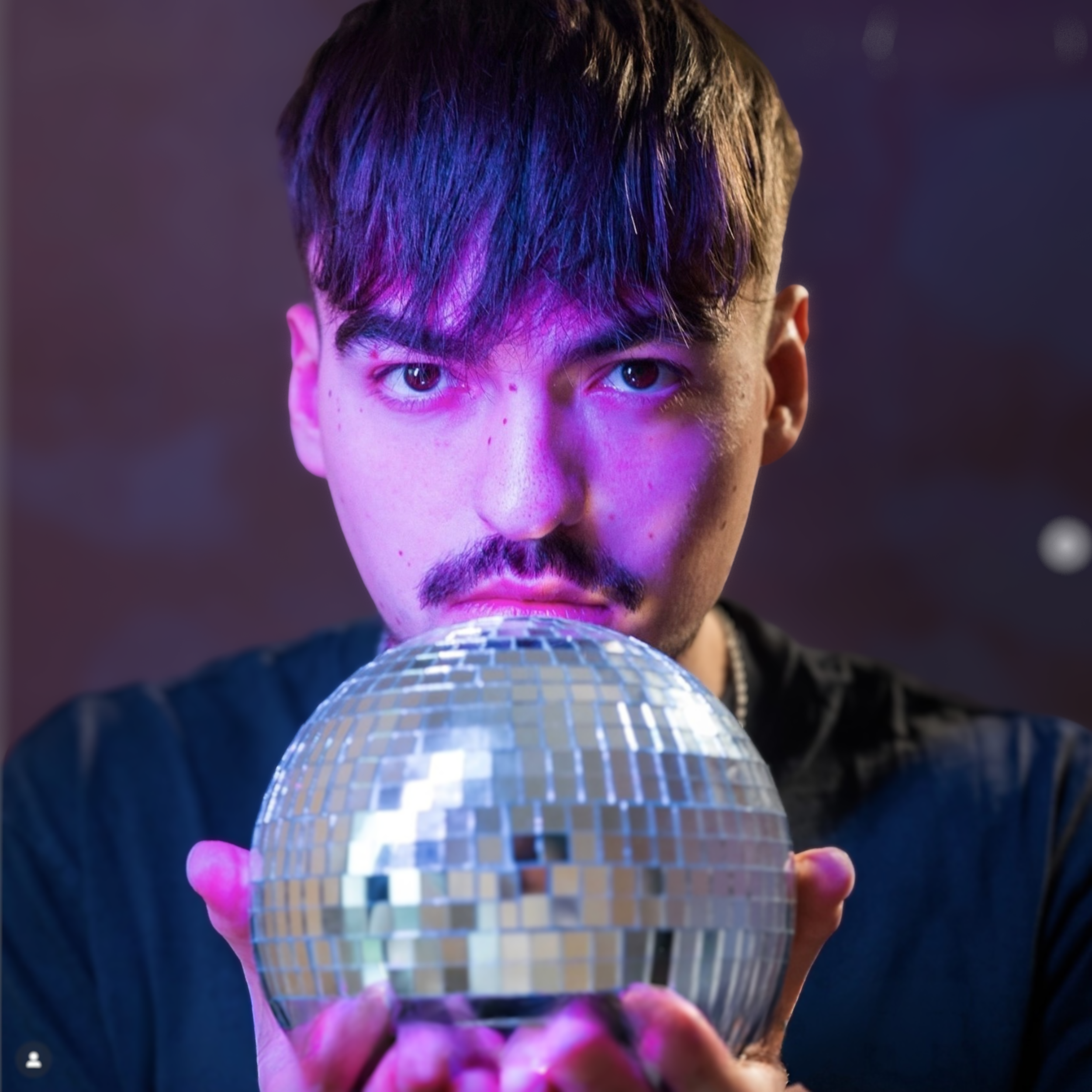 Junge Person mit dunklem Haar und Mustache hält einen kleinen Spiegelball, Blick direkt auf die Kamera gerichtet, mit violettem Licht beleuchtet.
