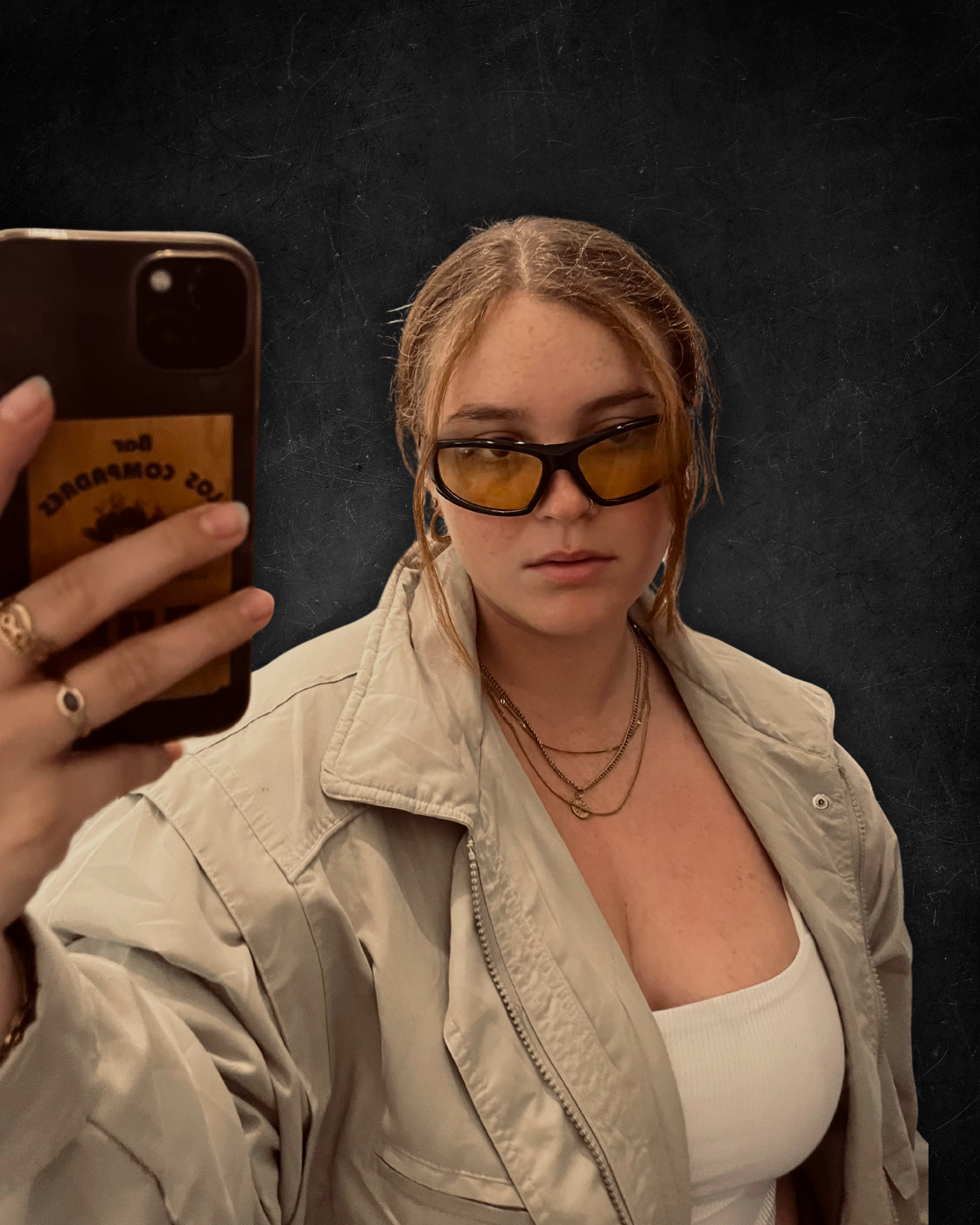 Junge Frau mit roten Haaren, schwarze Brille mit gelben Gläsern, beige Jacke, weiße Oberteil, in Schwarz gehaltenem Raum, macht Selfie mit Smartphone.