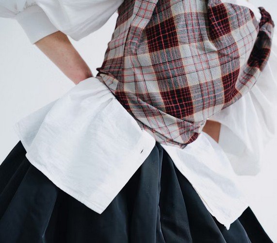 Person trägt einen weißen Hemd, einen schwarzen Rock und ein kariertes Tuch oder Schal.