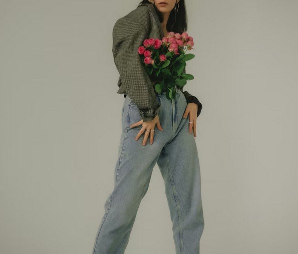 Eine Frau hält einen Bund pinker Rosen vor ihrem Oberkörper, trägt eine graue Jacke und weite Jeans, mit einer neutralen Hintergrund