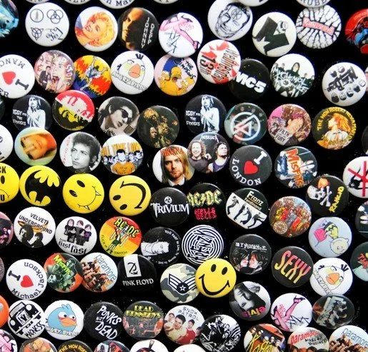 Sammlung von gedrückten Button-Buttons mit verschiedenen Designs, Logos, Emojis, Musikkünstlern und Marken auf einem schwarzen Hintergrund.