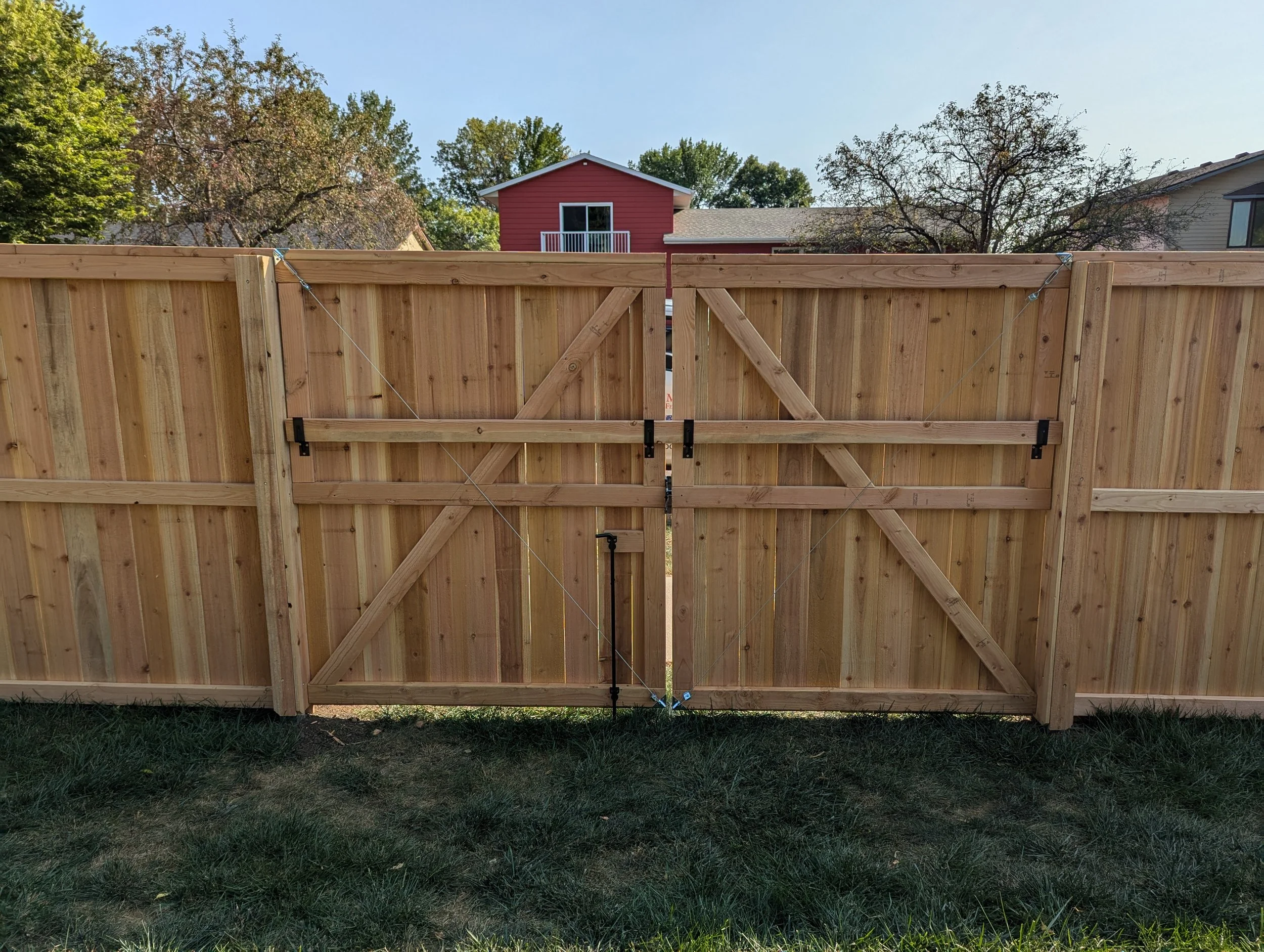 Newly installed wooden backyard fence with a double gate, metal hinges, and support cables, with trees and houses in the background under a clear sky.