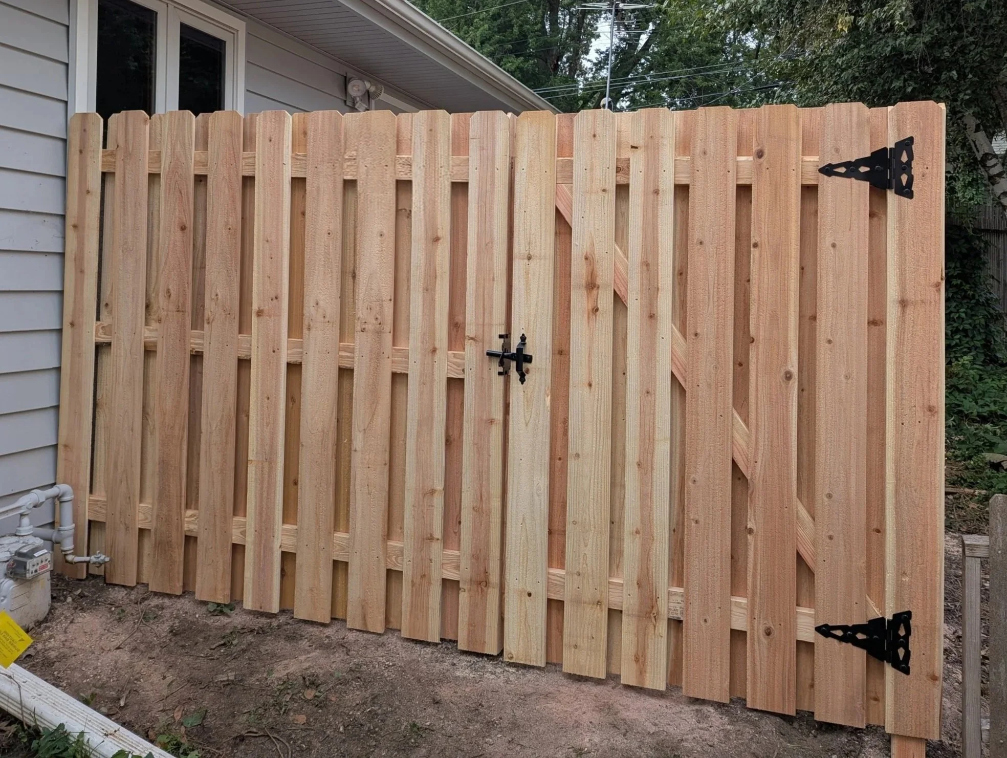 Finished wooden fence gate with black hinges and latch installed outside a house, with plants and trees in the background.