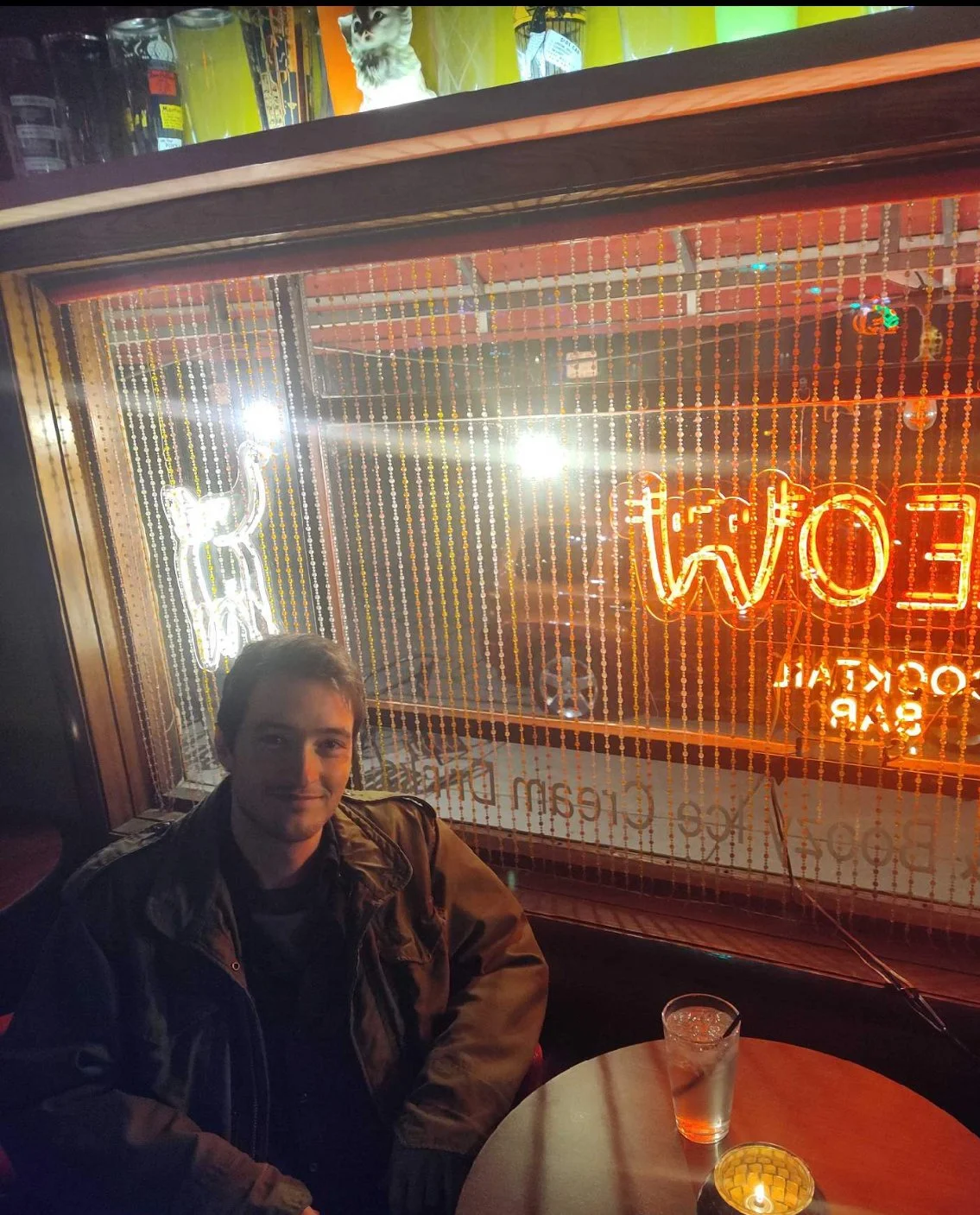A young man sitting at a round table inside a bar with neon signs and beaded curtains in the background. There is a glass with a drink and a lit candle on the table. Neon signs read 'OPEN' and 'LOS ANGELES'.