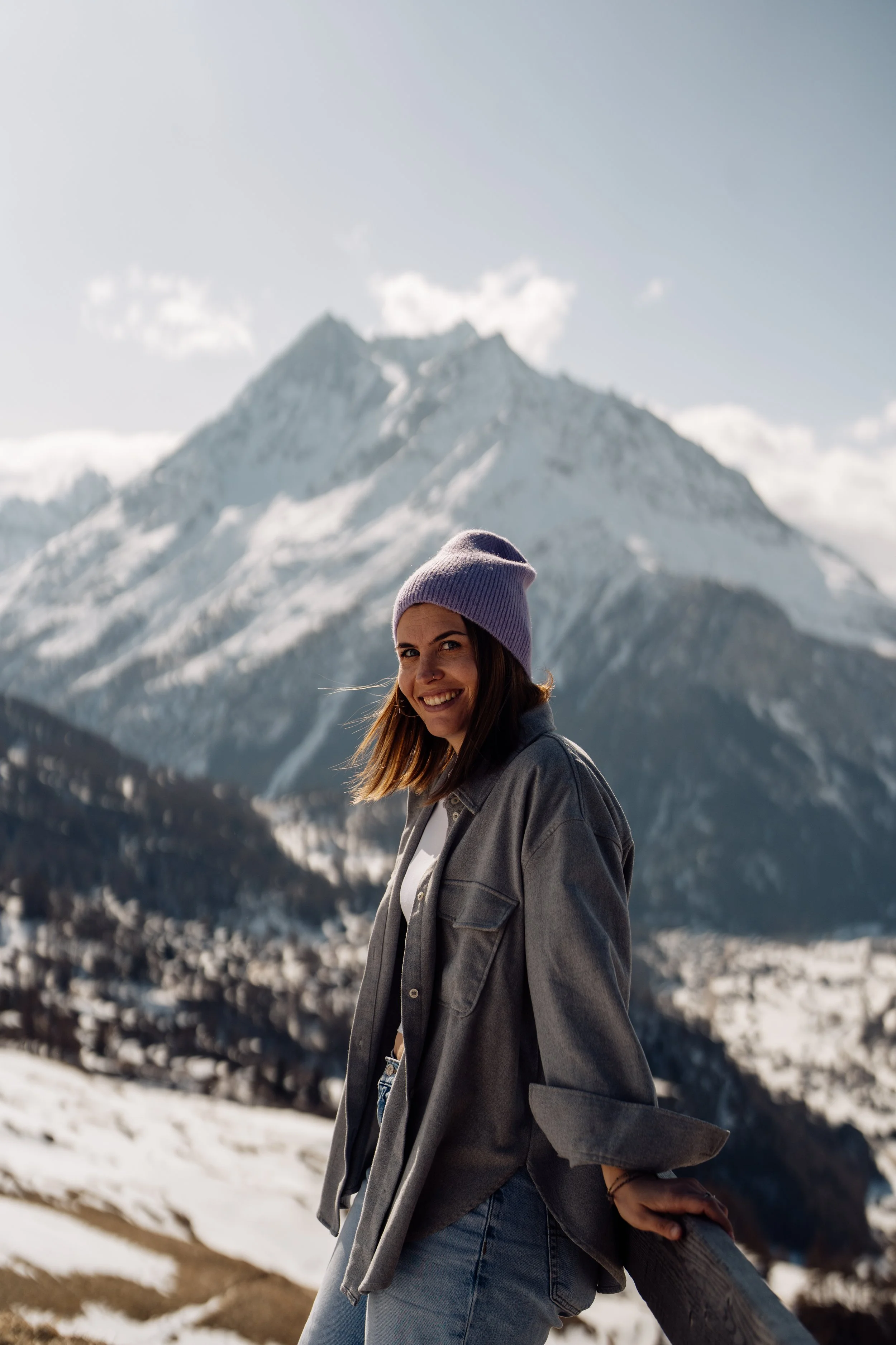Femme souriante en extérieur dans un paysage montagneux enneigé, portant un bonnet violet et une veste en denim gris.