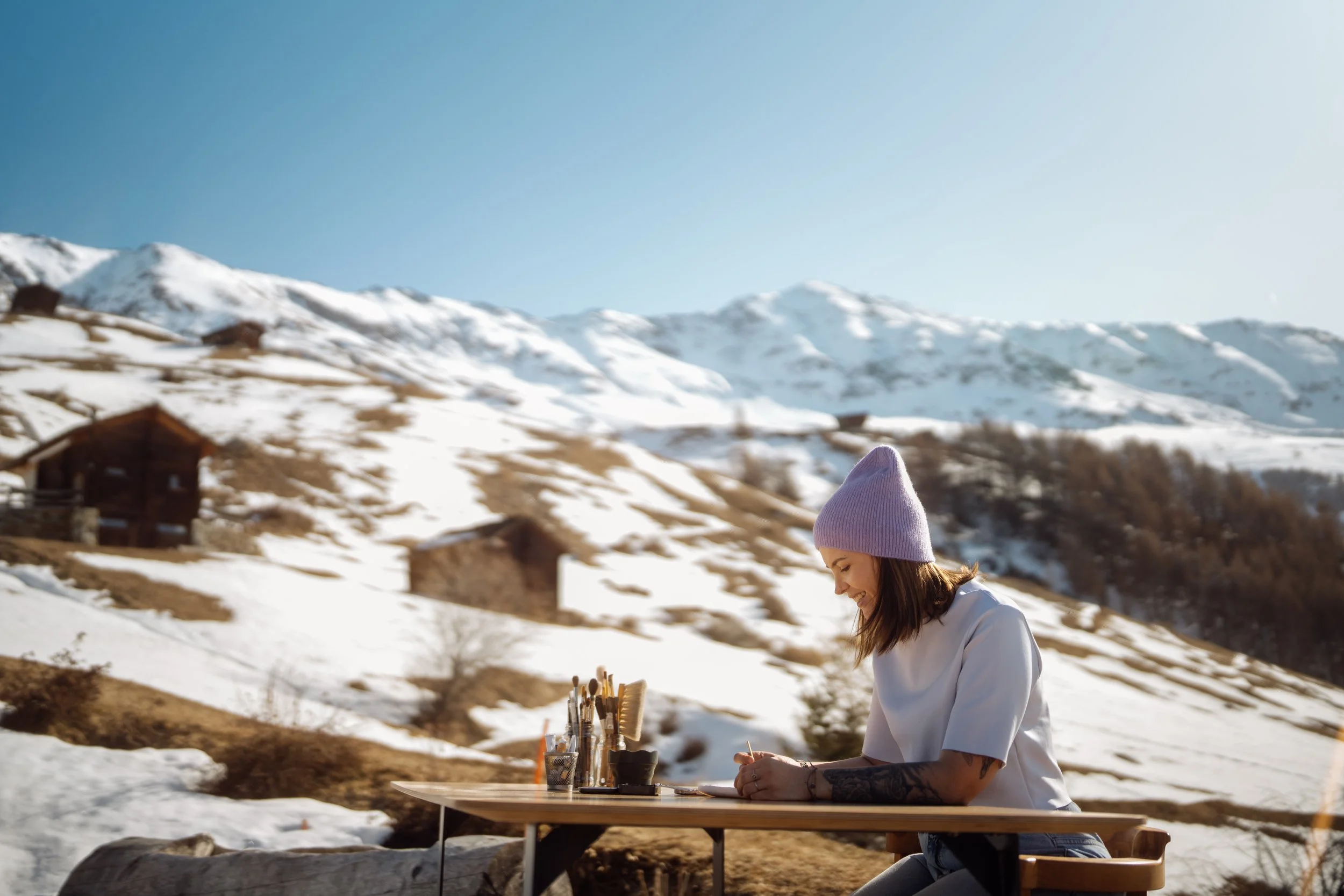 Femme assise à une table en plein air dans un paysage enneigé, portant un bonnet violet, écrivant ou dessinant, avec des montagnes enneigées en arrière-plan.