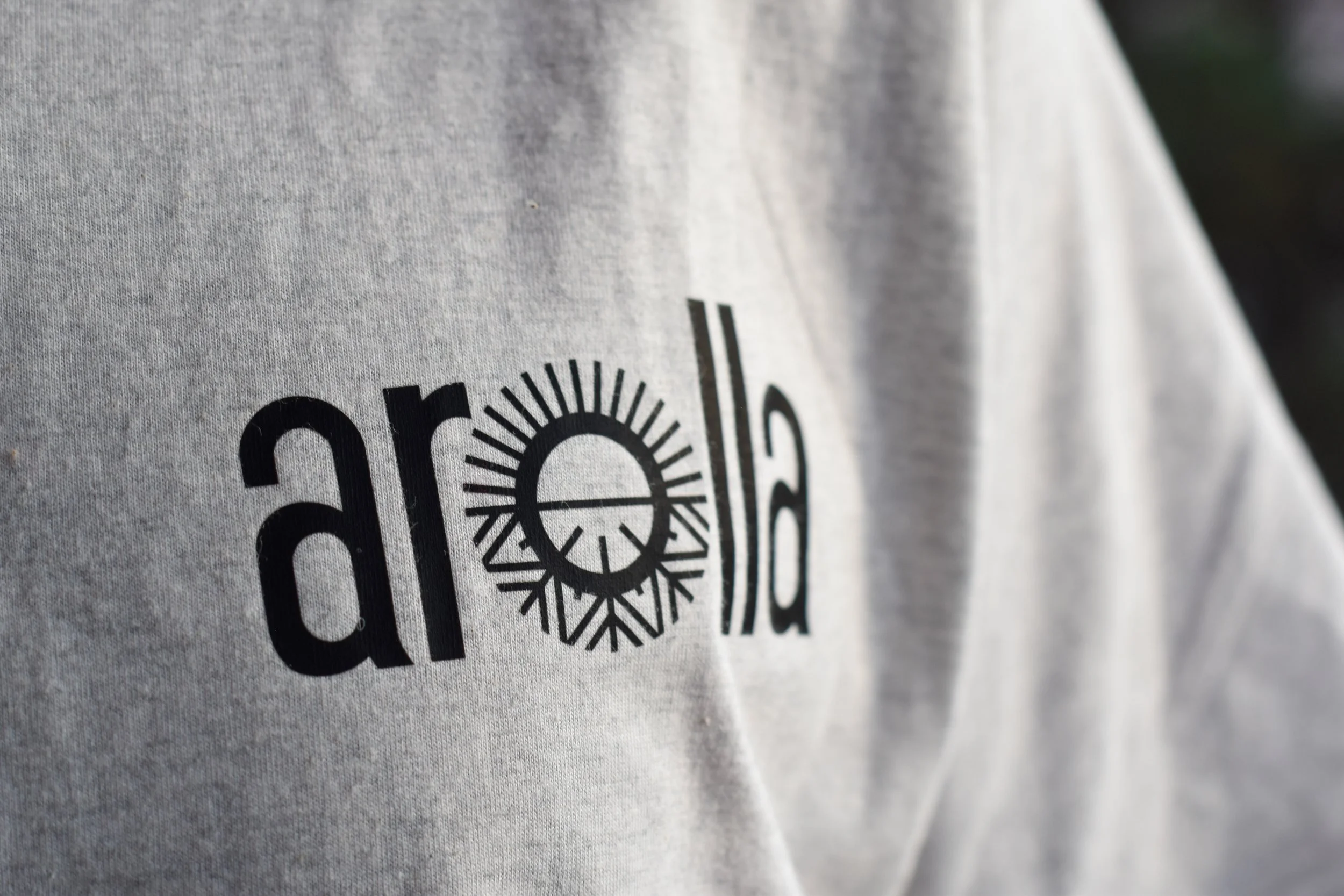 Zoom sur un t-shirt gris portant un logo noir avec le mot 'anold' stylisé, où le 'o' est remplacé par un cercle avec des rayons comme un soleil ou une roue, et une ligne horizontale traversant le cercle.