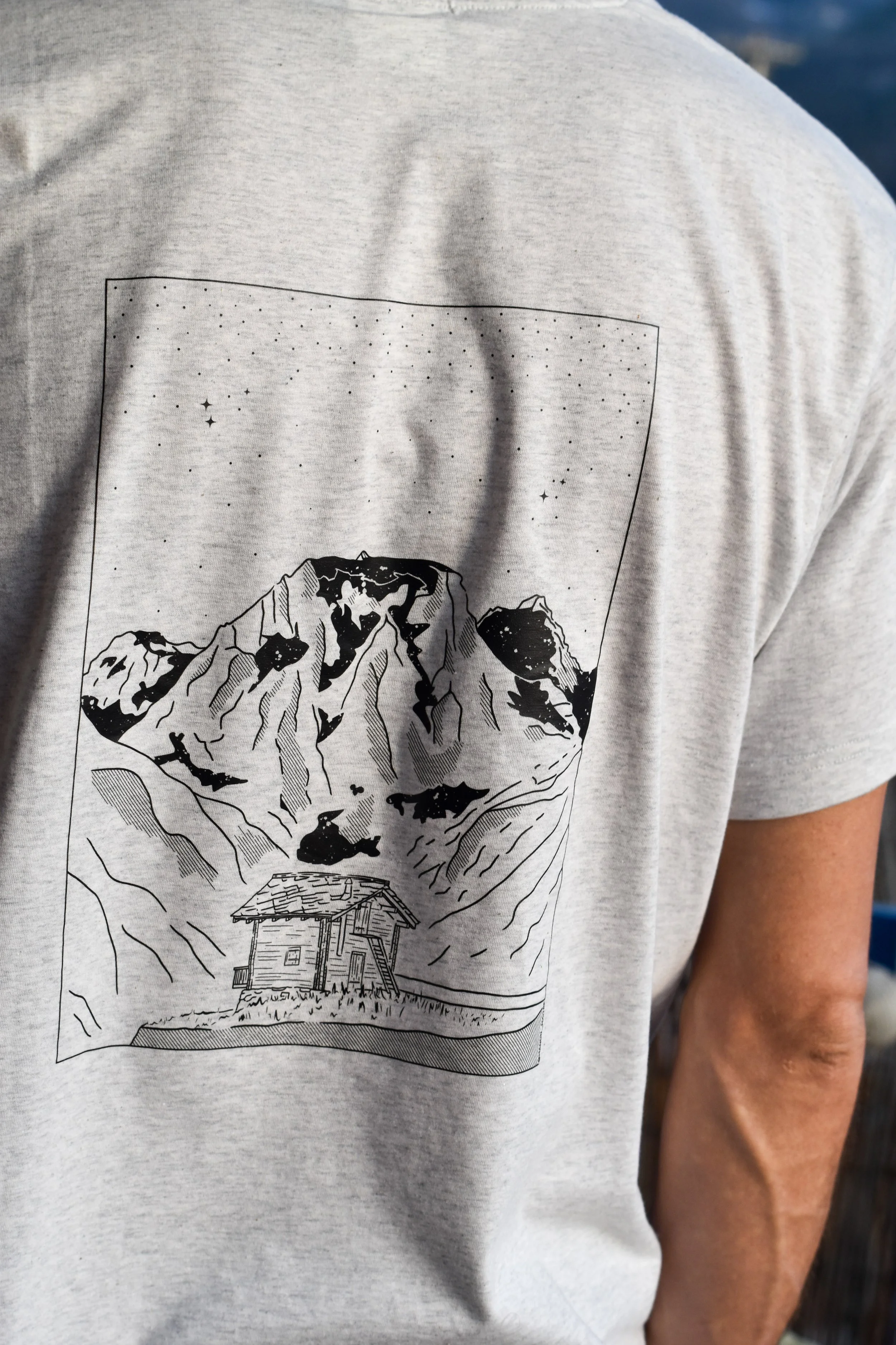 T-shirt gris avec illustration d'une maison en montagne, grands sommets enneigés et ciel étoilé.