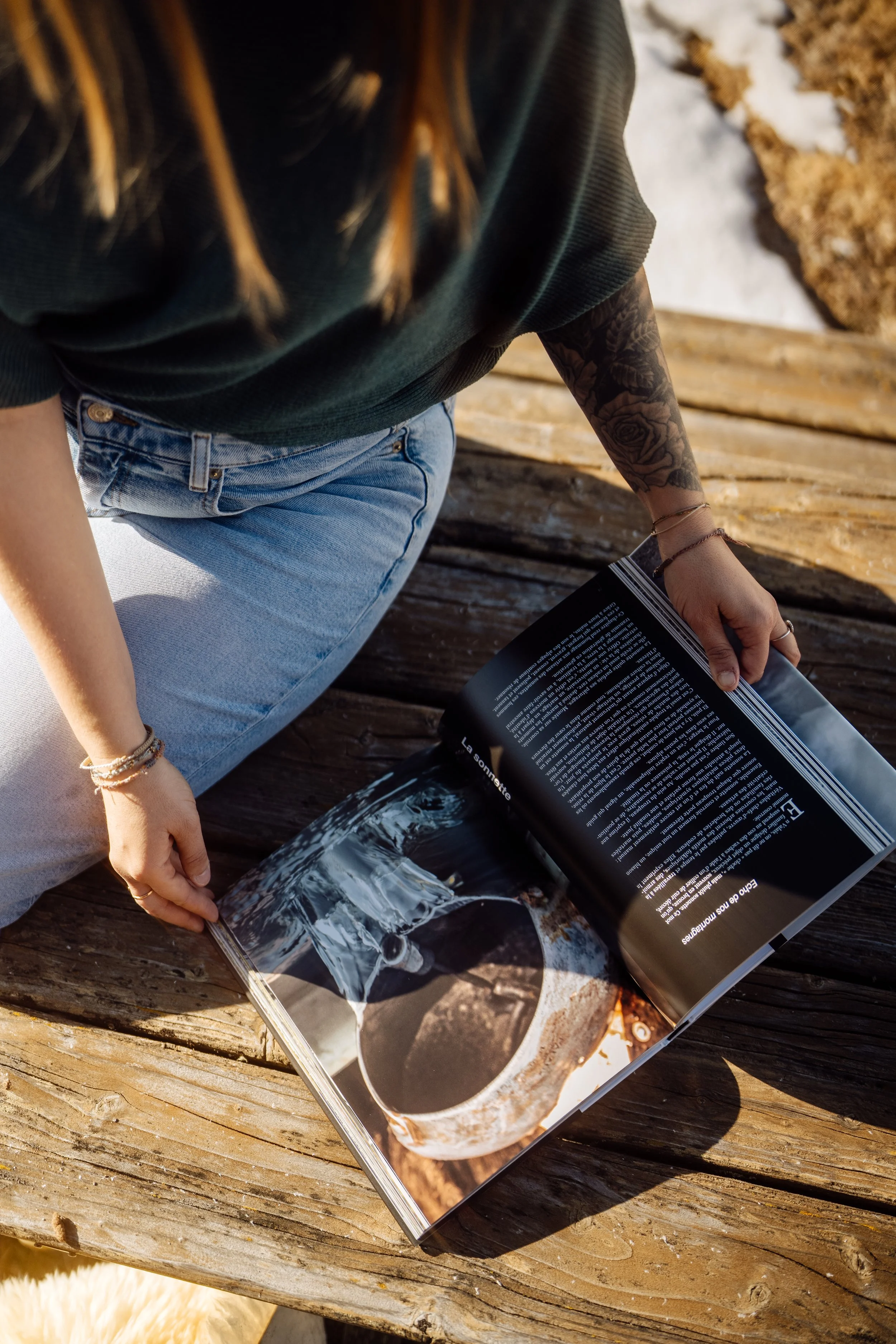 Personne assise sur un banc en bois, lisant un magazine avec une photo d'une tasse de café en gros plan. La personne porte un t-shirt vert, un jean et a des tatouages sur le bras. La scène est prise en extérieur, avec un sol partiellement enneigé en arrière-plan.
