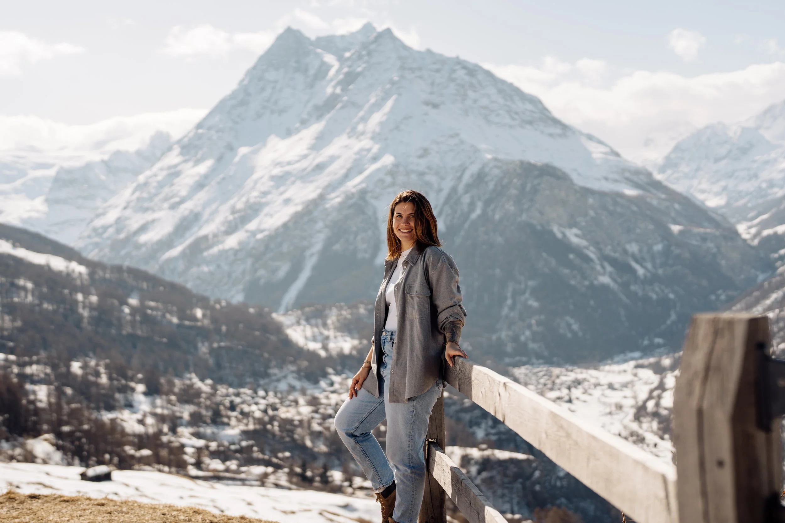 Femme souriante debout près d'une barrière en bois avec des montagnes enneigées en arrière-plan.