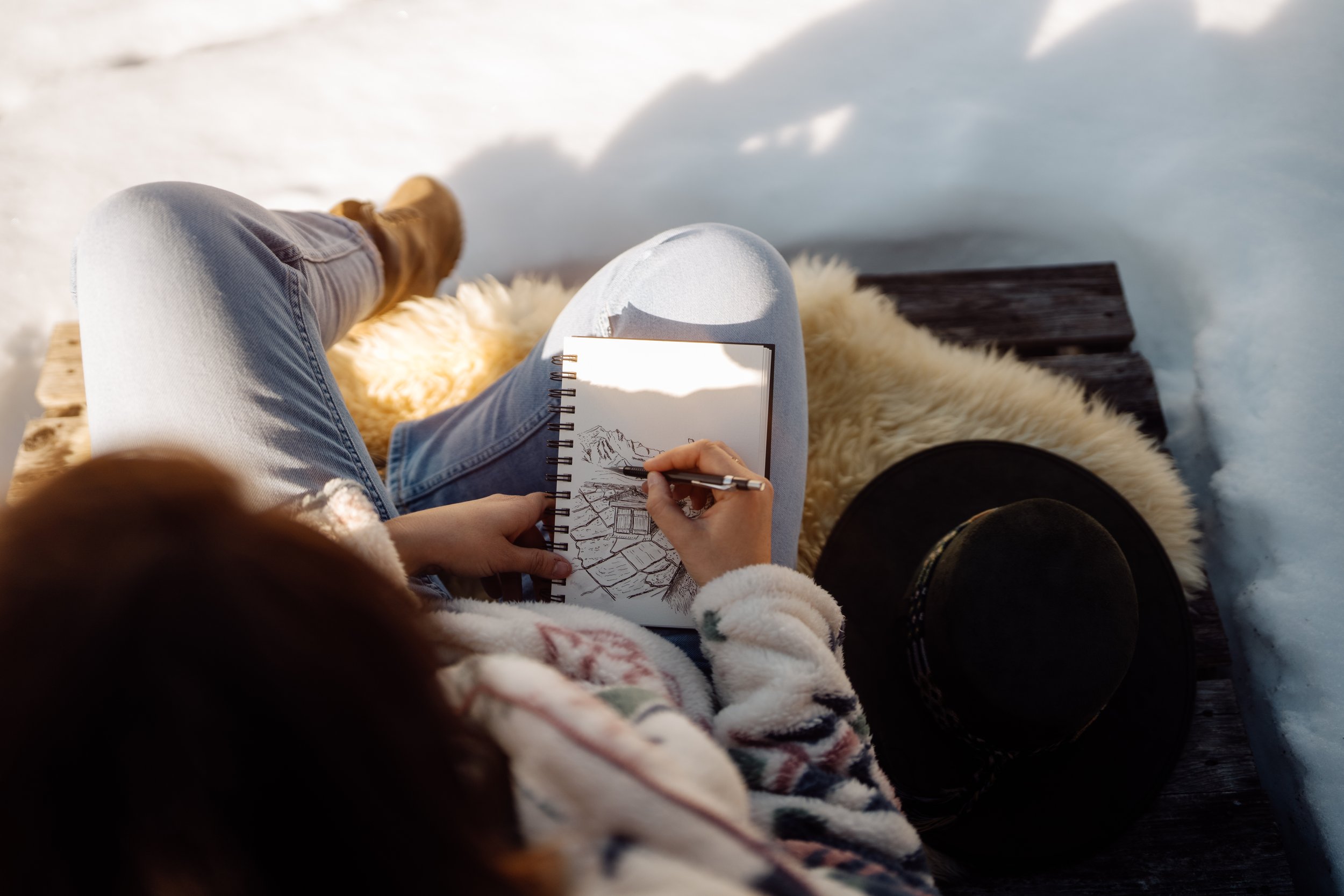 Personne assise sur un banc en bois dans la neige, dessinant un paysage de montagnes dans un carnet, avec un chapeau noir posé à côté sur le banc, portant un manteau en fourrure et des bottes en cuir.
