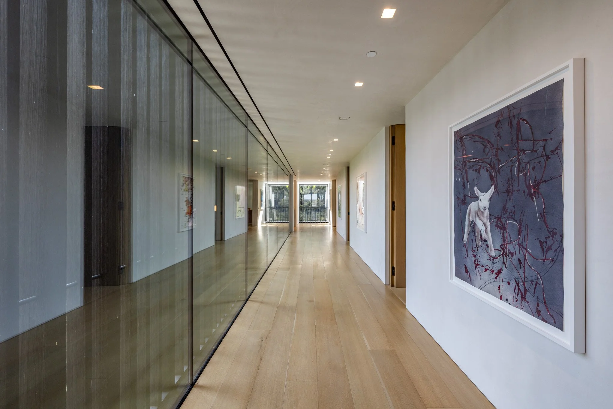 Modern art gallery hallway with white walls, wooden flooring, glass wall on the left, and framed artwork, including a painting of a white dog, on the right.