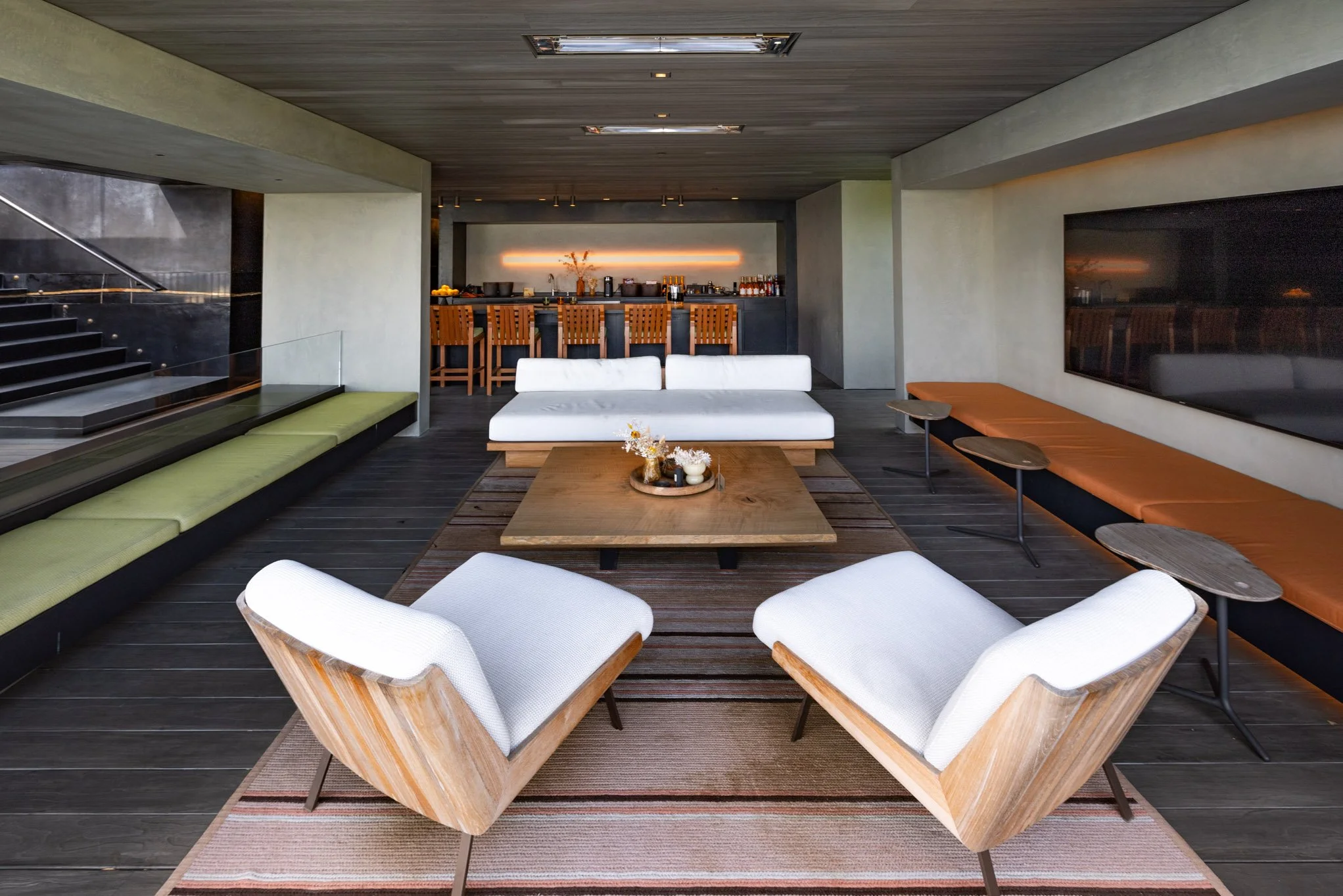 Modern living room with white armchairs, wooden coffee table, and long cushioned bench, with a bar in the background.