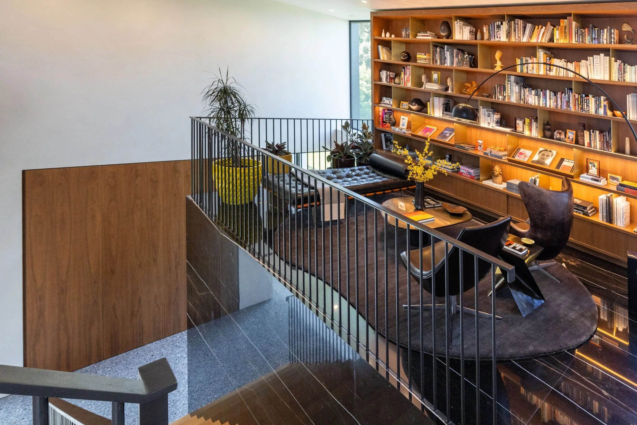 A cozy reading nook with a large wooden bookshelf filled with books, a black leather bench, flowers in a vase, and comfortable chairs on a dark rug, next to a large window with natural light.
