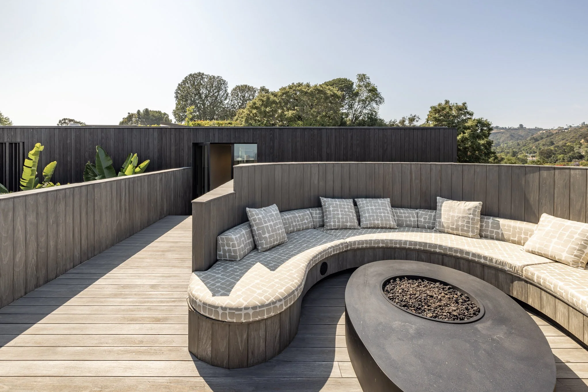Outdoor rooftop lounge with curved built-in beige cushion seating with patterned pillows, a black oval fire pit, wooden deck flooring, and modern black and gray walls with lush green trees in the background.