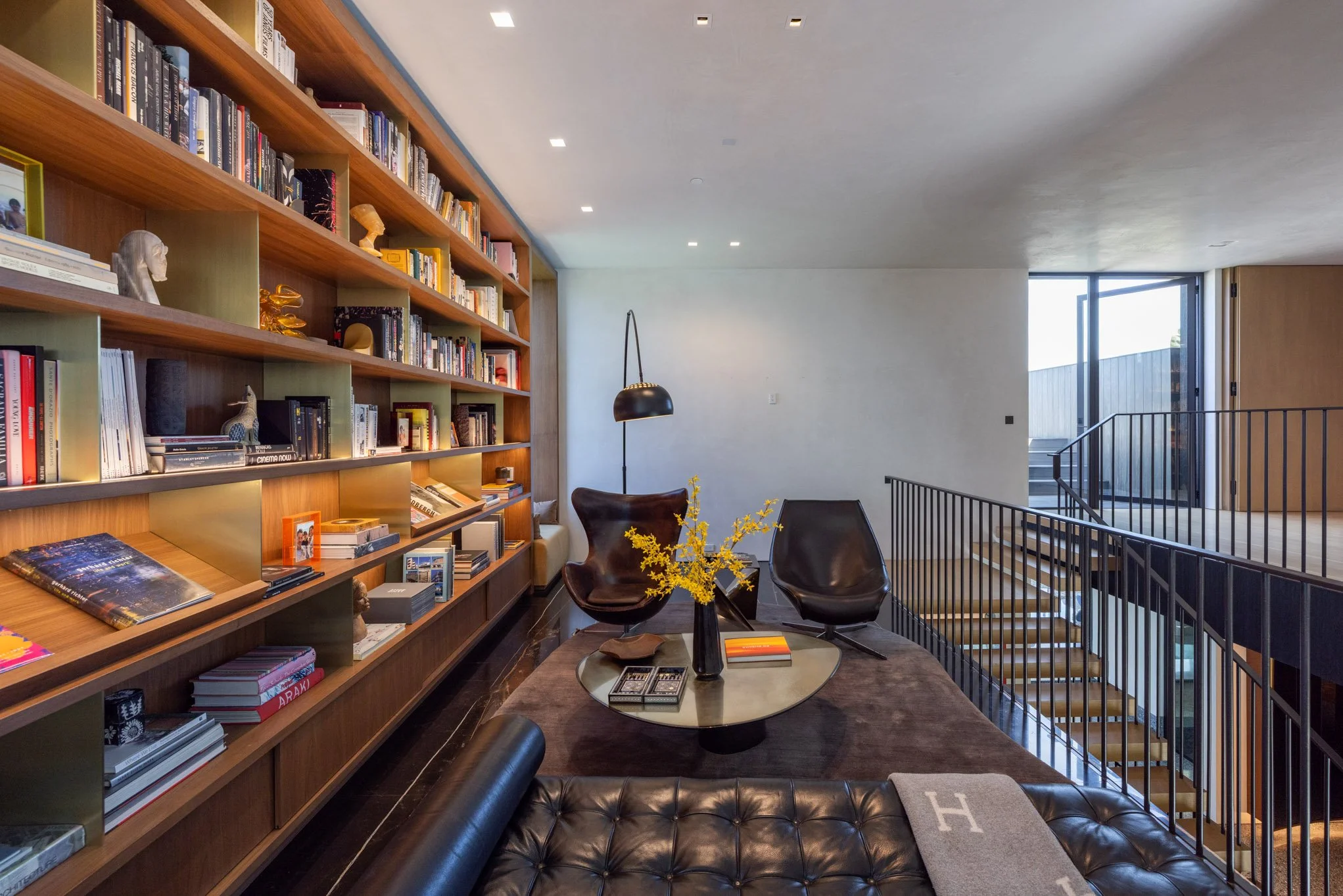 Modern living room with a large wooden bookshelf filled with books and decorative items, a glass coffee table with magazines and a vase with yellow flowers, black leather chairs, a floor lamp, a leather couch with a throw blanket, and a staircase wit