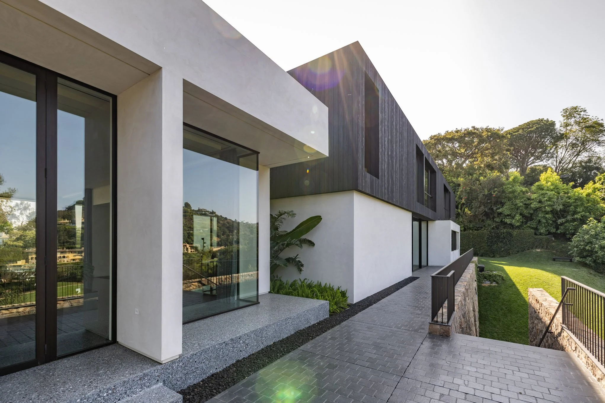 Modern residential building with large glass windows, white and dark wood exterior, situated on a landscaped hill with greenery and trees.