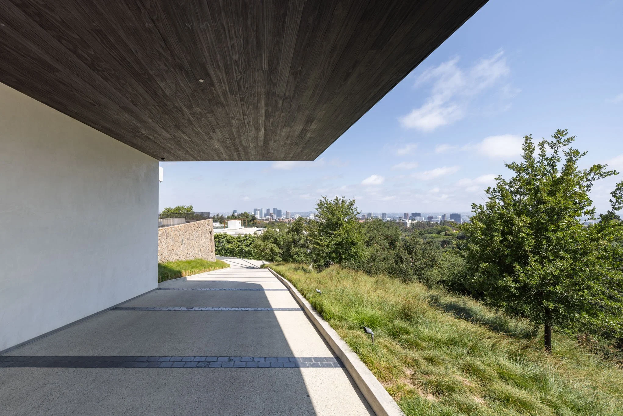 Modern building exterior with overhanging roof, paved walkway, and view of city skyline and trees in the distance.