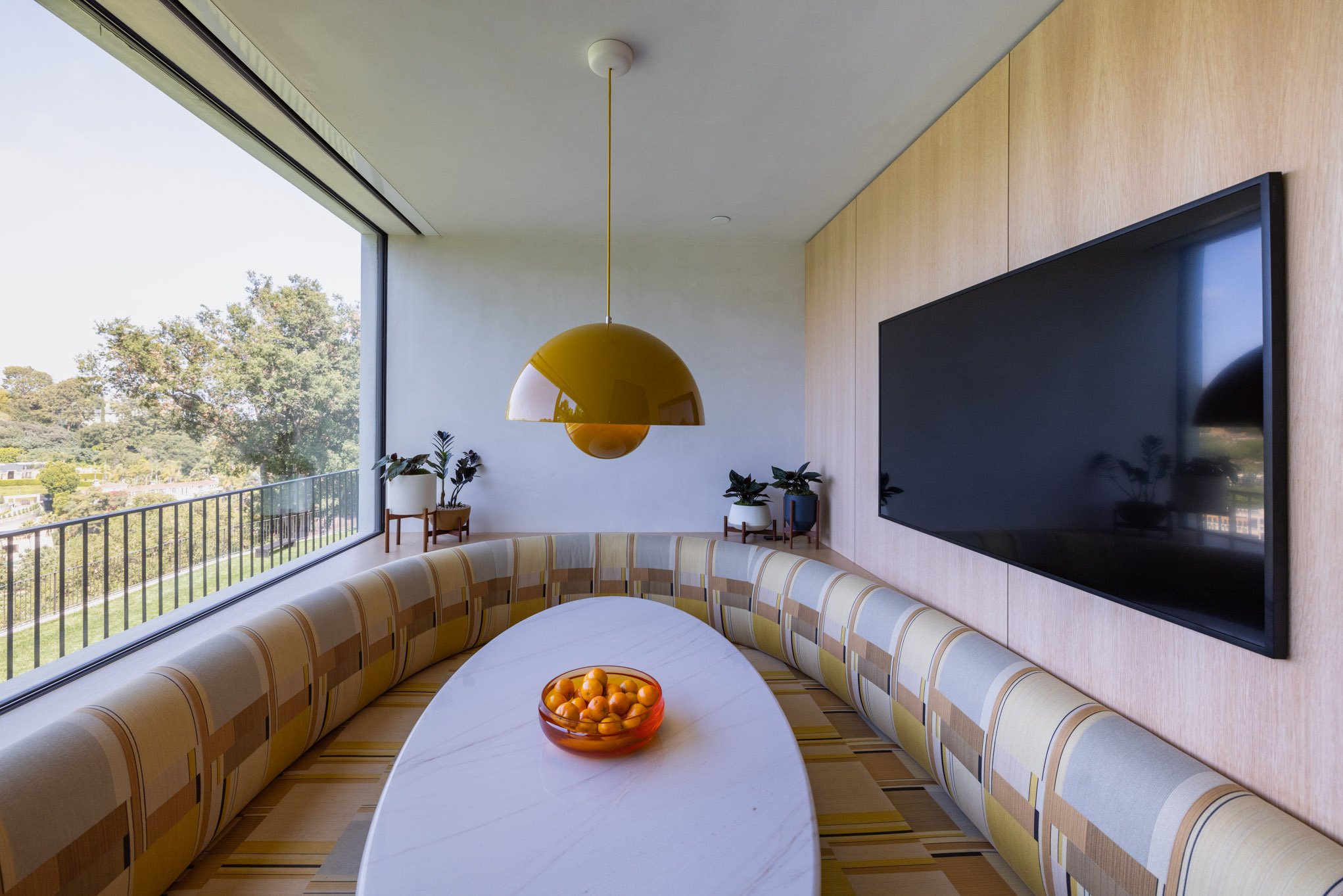 Modern dining area with a curved bench, white marble table with a bowl of tangerines, large window showing outdoor greenery, wooden wall with a flat-screen TV, and a yellow pendant light.