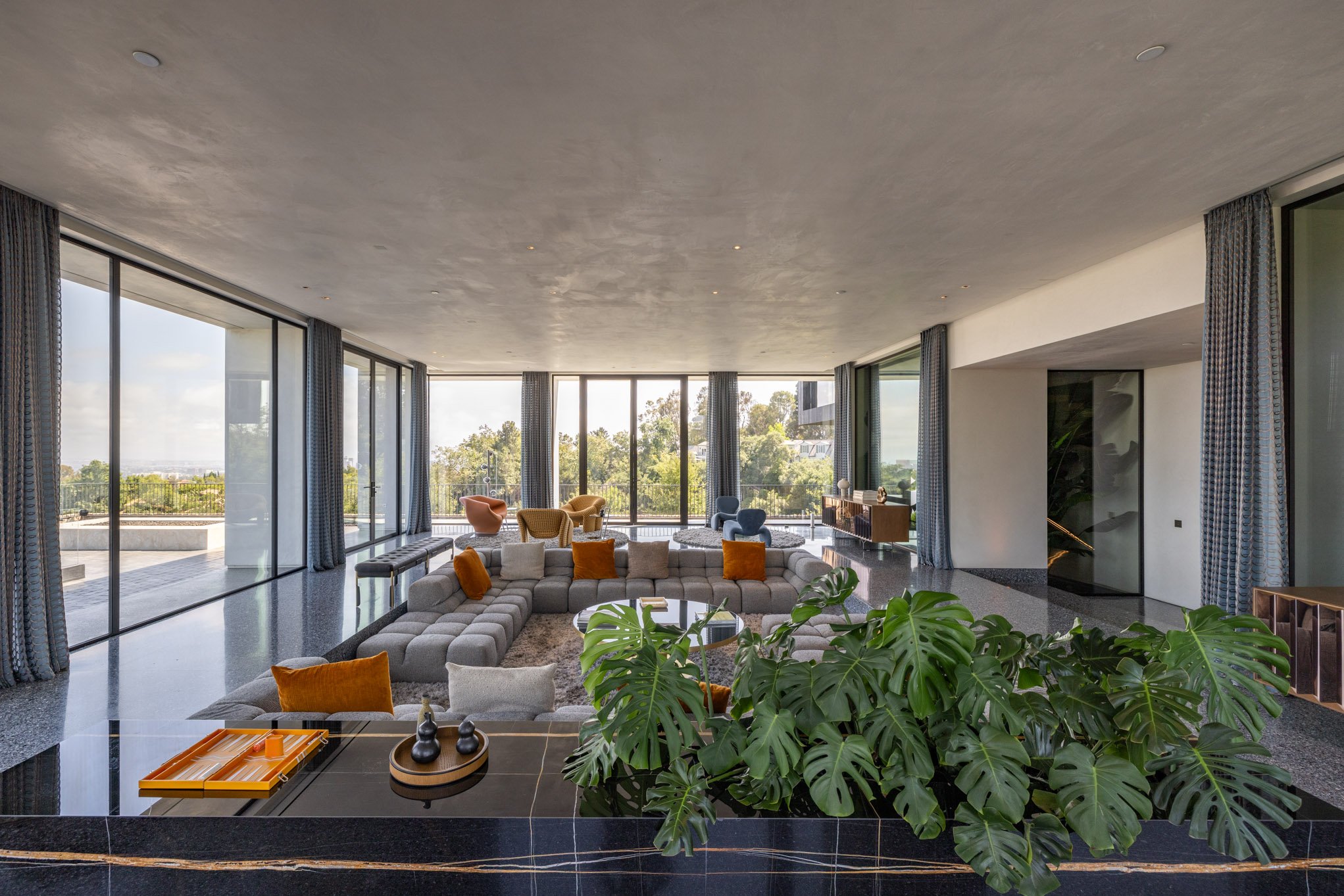 Modern living room with large windows, beige sectional sofa with orange and beige pillows, blue and peach armchairs, a coffee table, and green plants, overlooking a patio and green landscape.