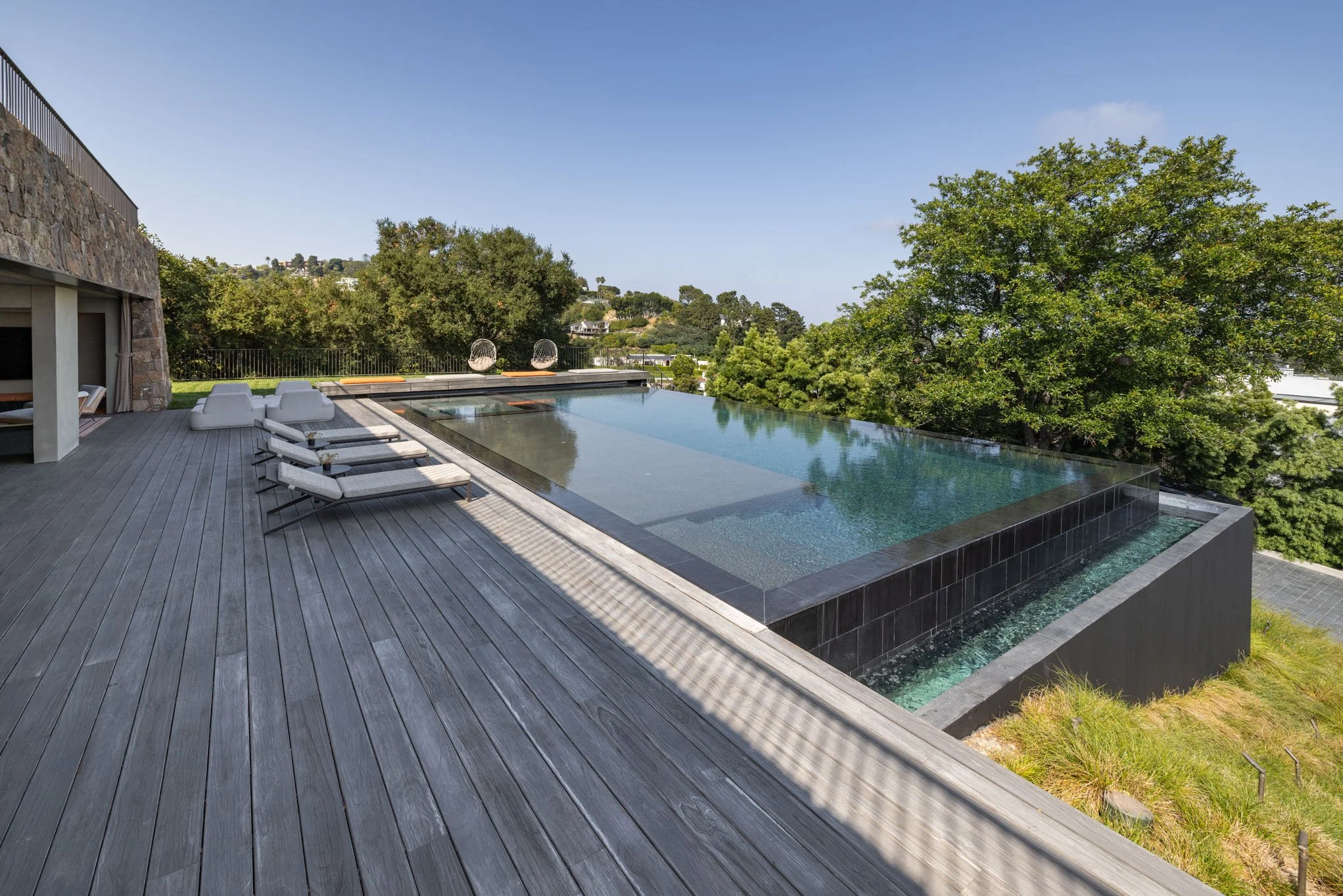Luxury rooftop infinity pool with lounge chairs on wooden deck, surrounded by lush green trees and clear blue sky.