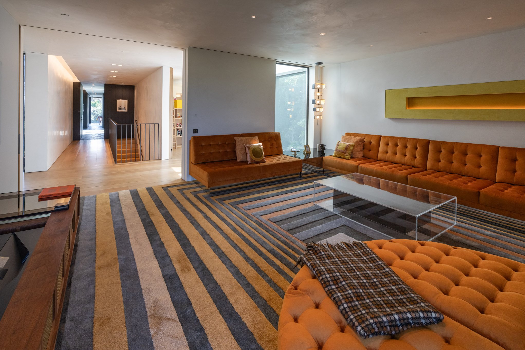 Modern living room with orange tufted sofas, a glass coffee table, striped rug, and large windows.