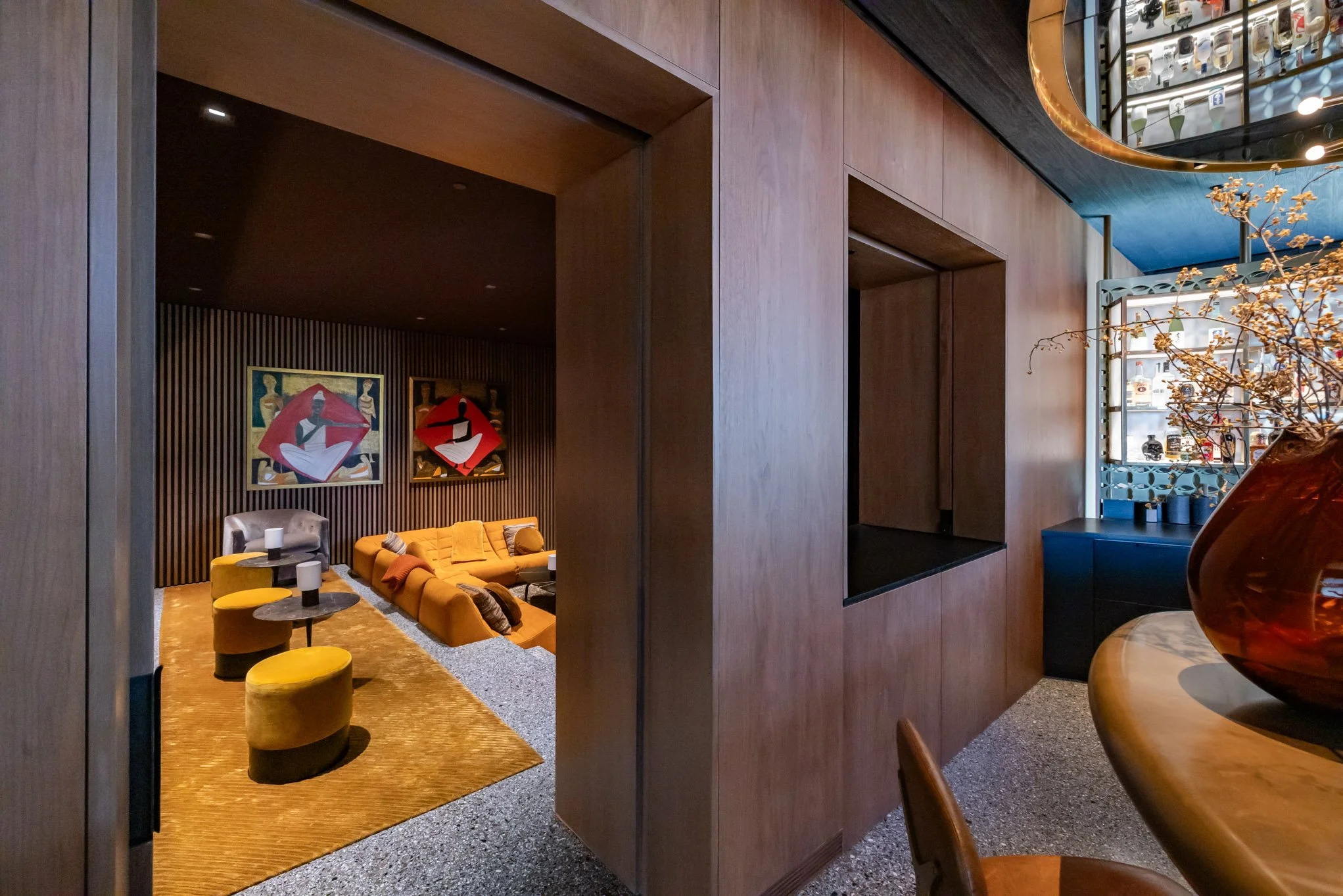 Interior of a modern, stylish lounge area with yellow and grey furniture, framed artwork on a black wood-paneled wall, and decorative vases and plants.