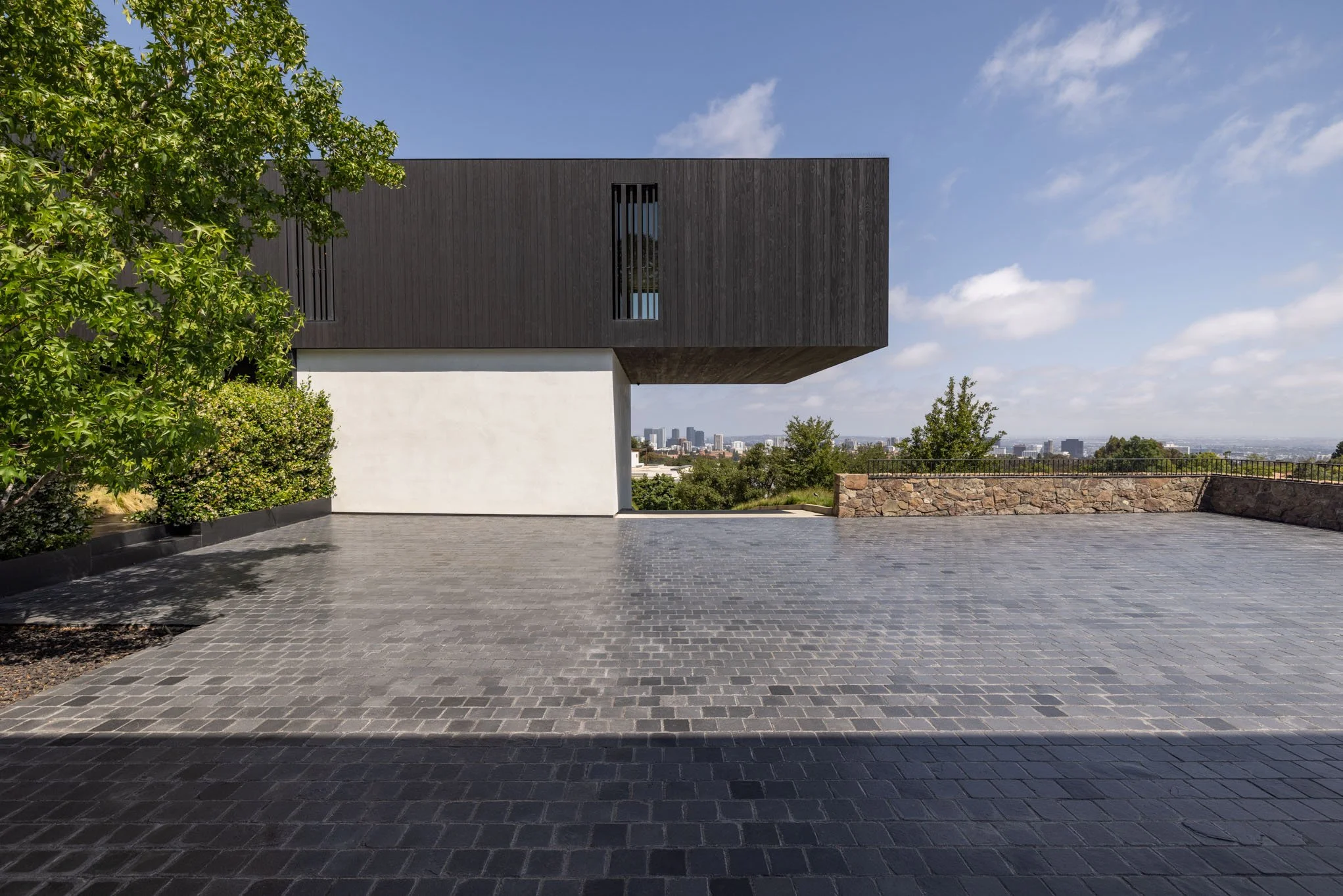 Modern building with dark and white exterior, surrounded by a paved terrace, greenery on the left, city skyline in the distance under a blue sky with scattered clouds.